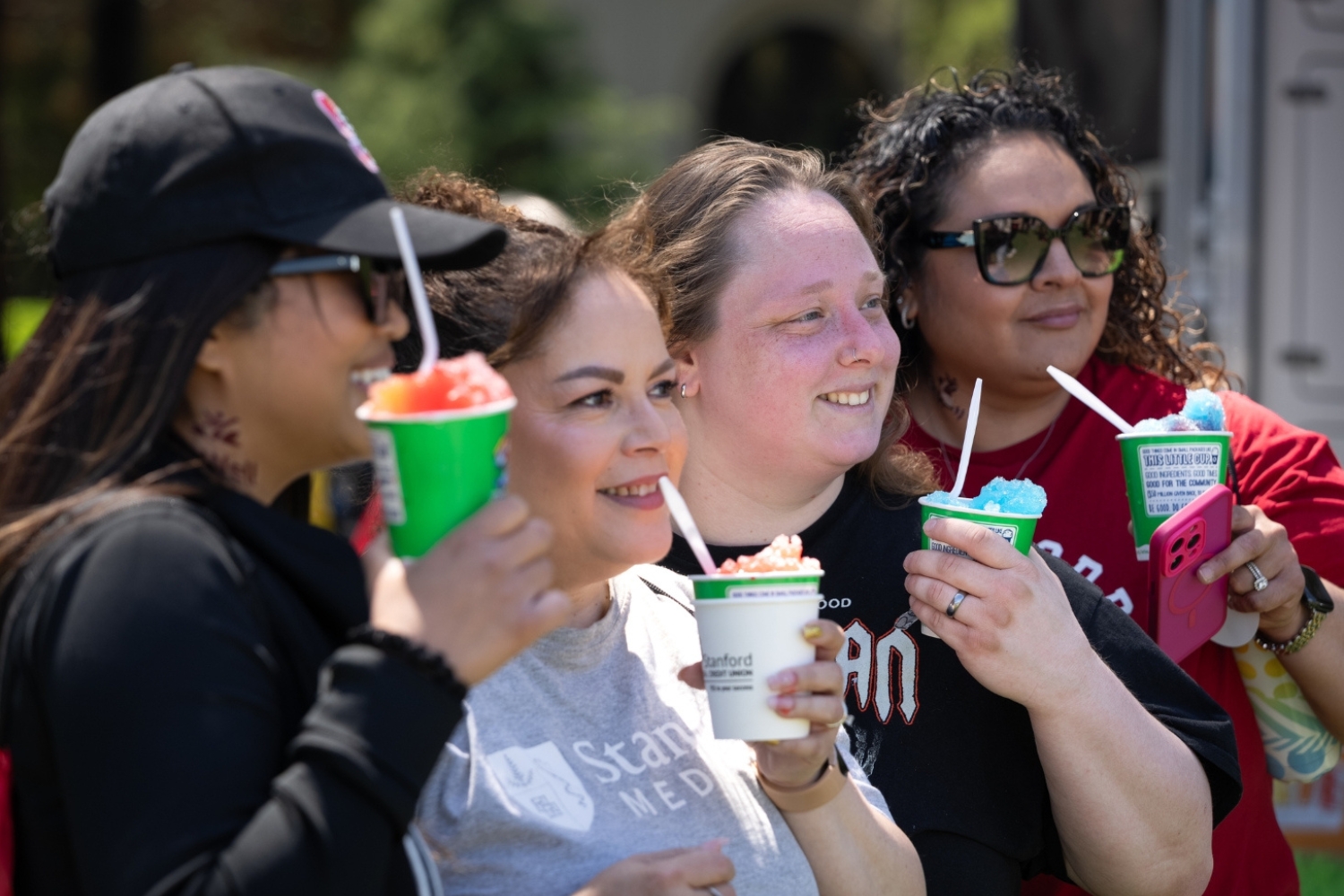 A close-up of friends enjoying colorful frozen treats in cups while smiling and chatting during an outdoor event.