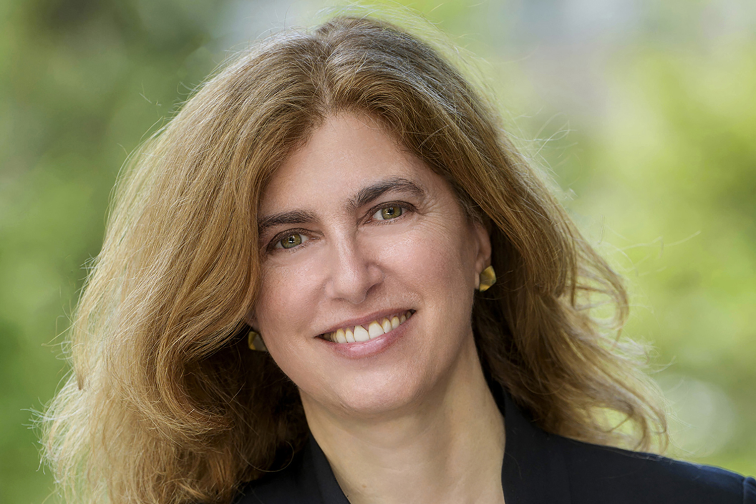 Closeup of Jessica Riskin with wavy, shoulder-length hair standing outdoors, wearing a black jacket and gold earrings against a blurred green background.