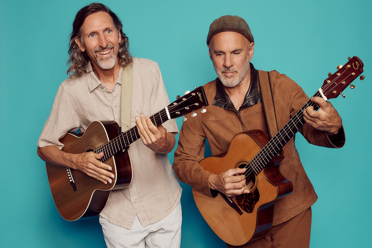 West and Strauss pose with acoustic guitars in front of a light blue background, showcasing their instruments and relaxed expressions.