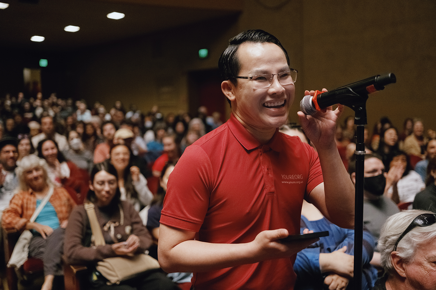 An audience member in a red shirt holds a smartphone while speaking into a microphone at an event filled with attendees.