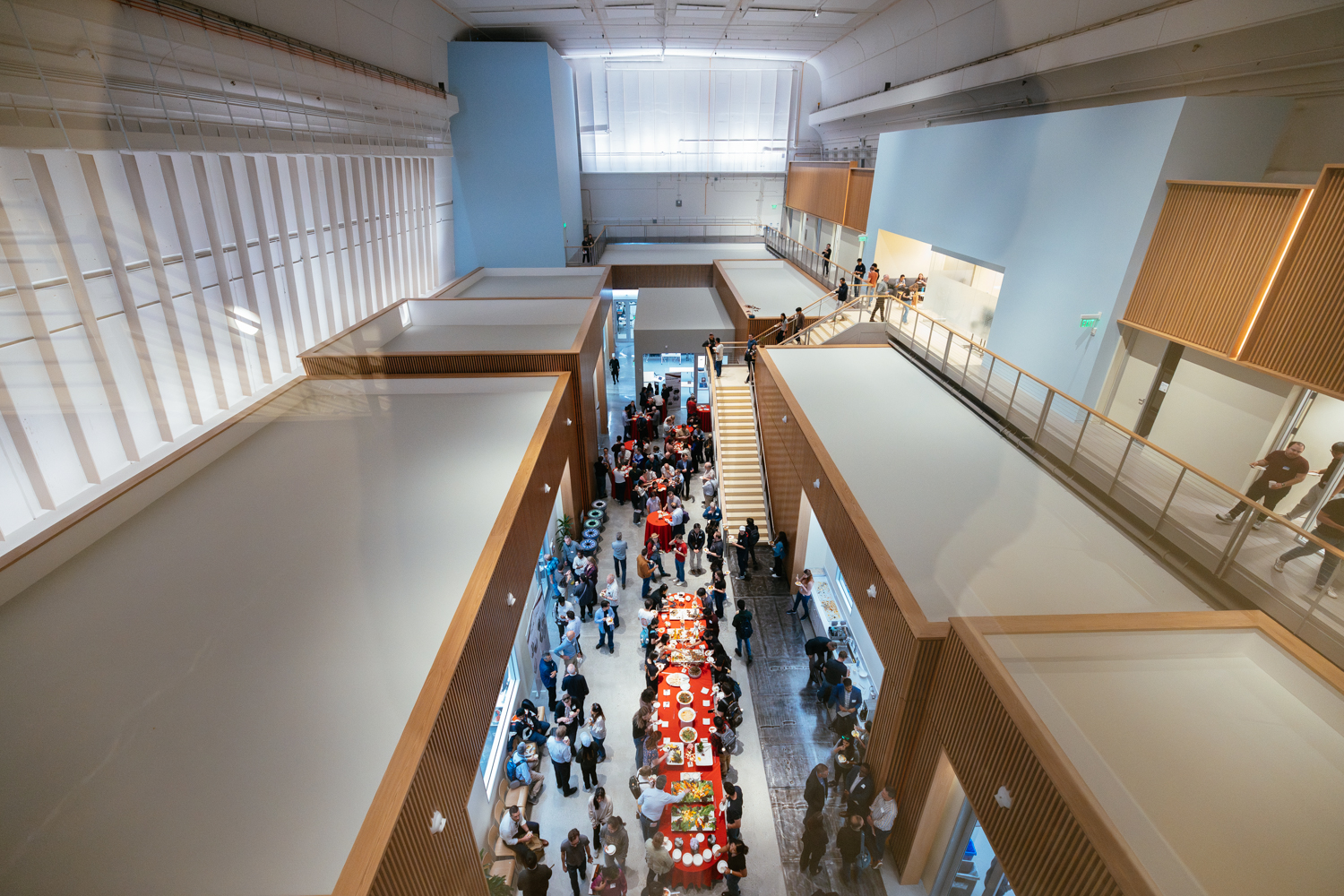 An aerial view of a modern event space filled with people mingling around a red table set with food.