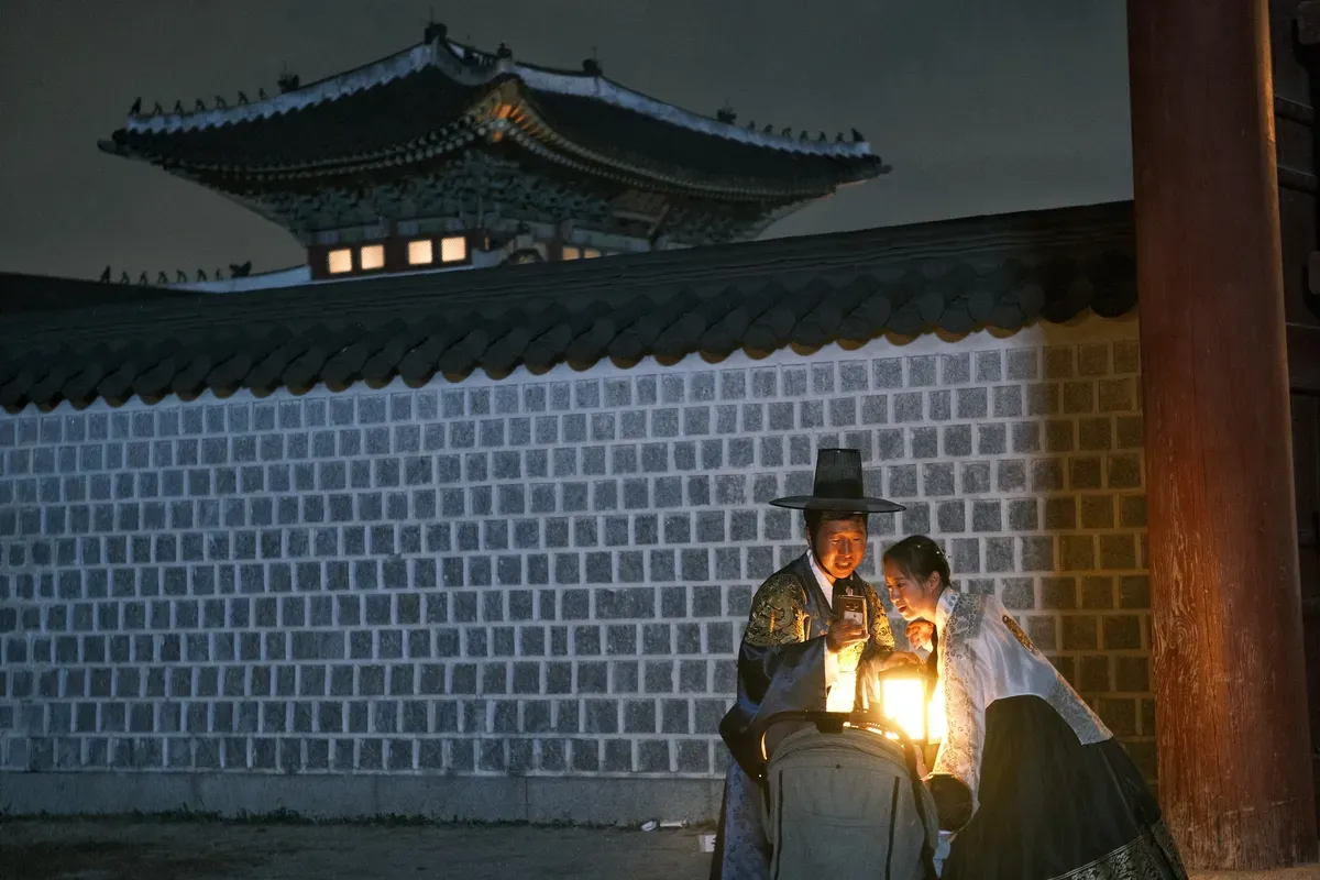 Man and woman in traditional Korean clothing examine a lantern near a historic building at night.