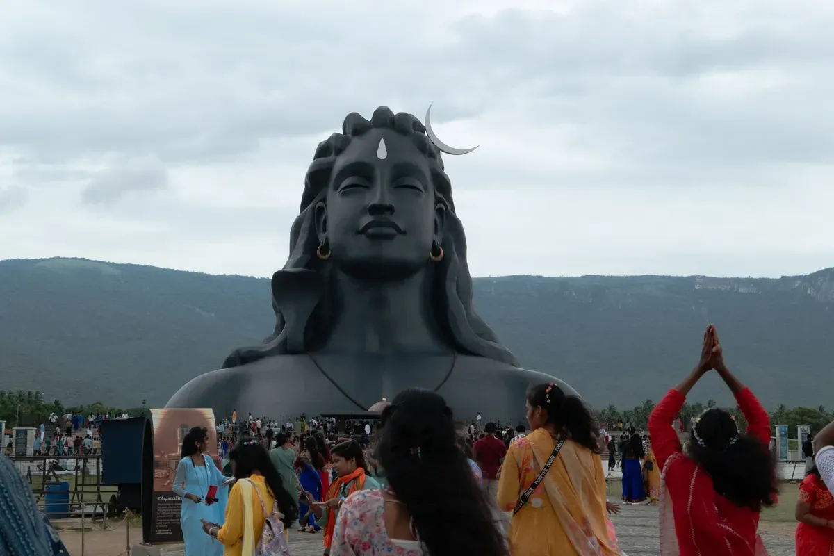 Visitors gather near a large statue of Shiva, with many individuals shown in prayer or reflection against a cloudy backdrop.