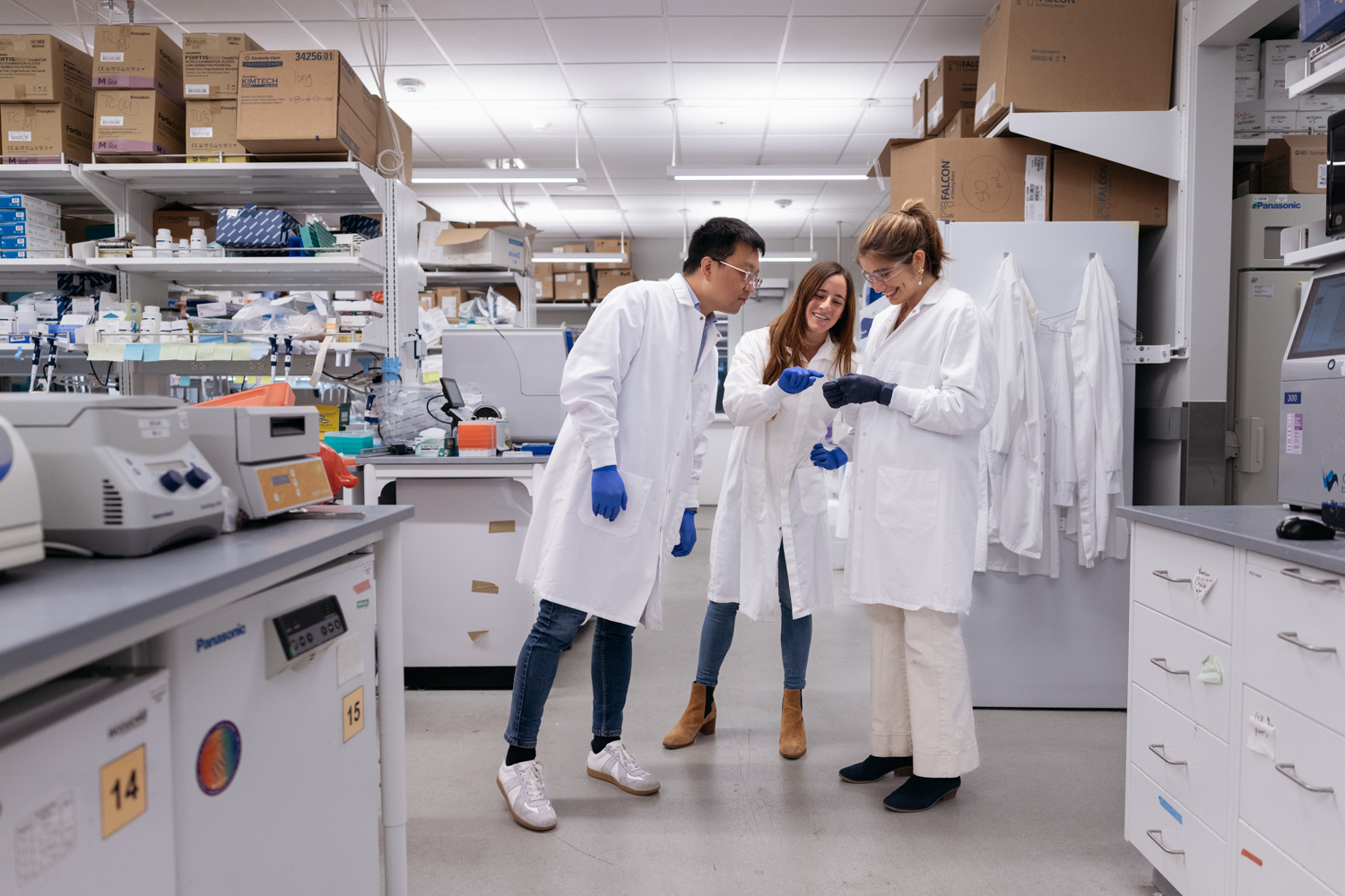 Researchers in lab coats and gloves discuss data while standing in a lab filled with equipment and supplies.