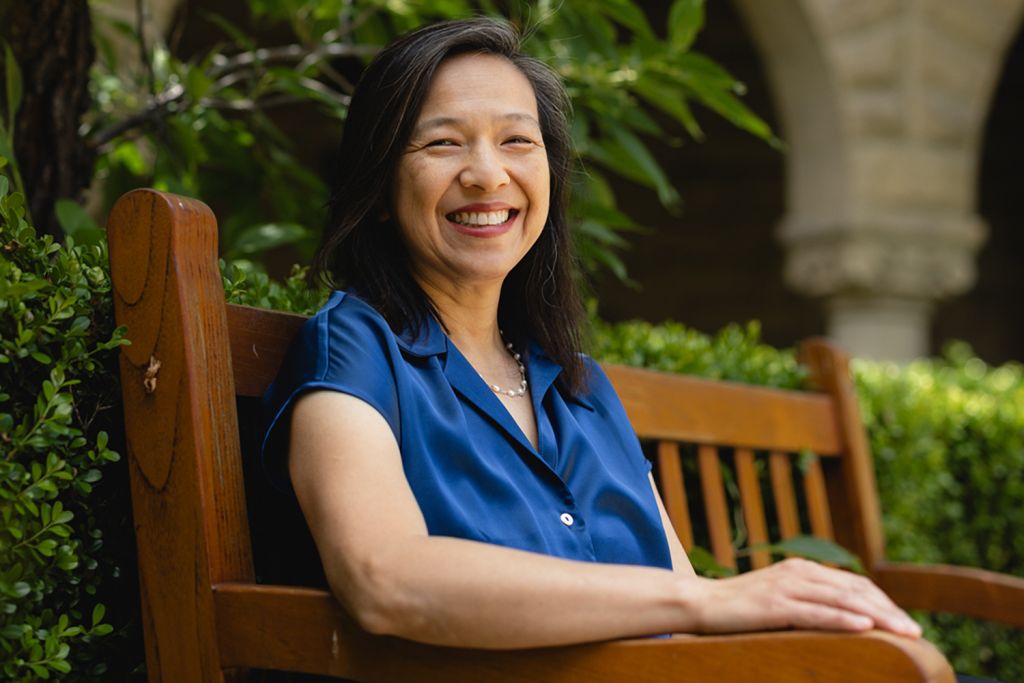 Mary Chen in blue short sleeve top smiling while sitting on a wooden bench surrounded by greenery.