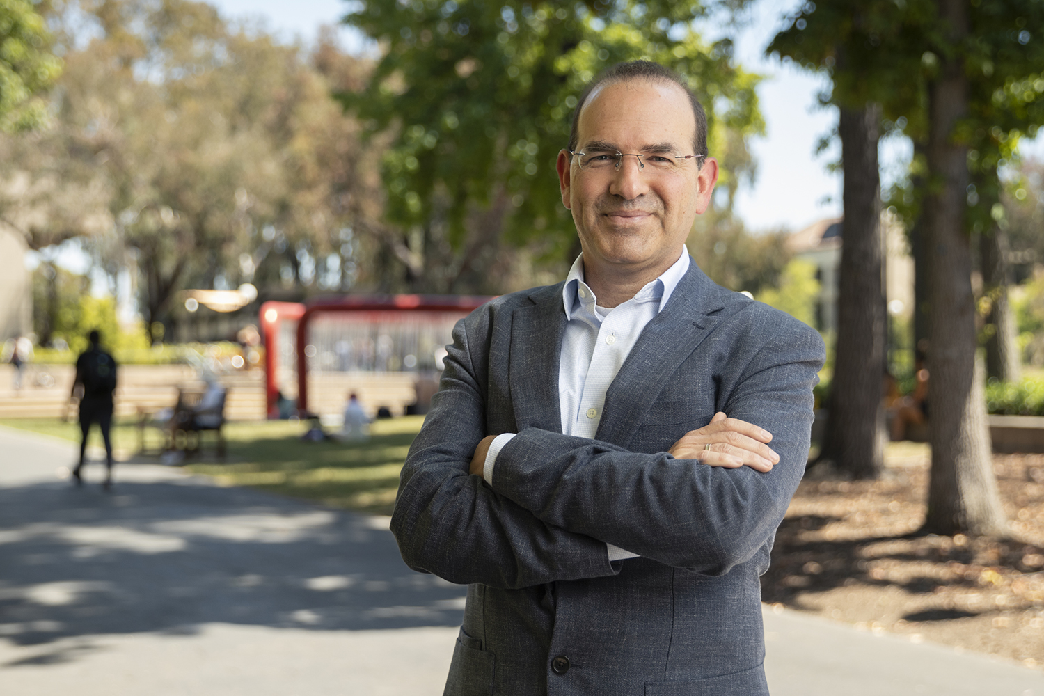 Abramitzky in gray suit and glasses standing with arms crossed on a tree-lined campus path with red hoop fountain in background.