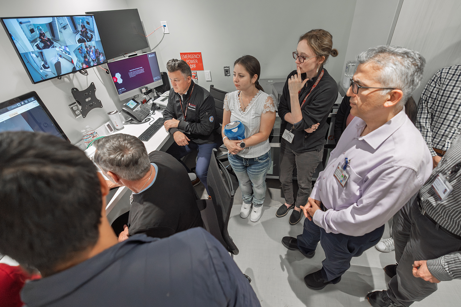 A group of people observes screens in a control room, focused on monitoring activities in a medical setting.
