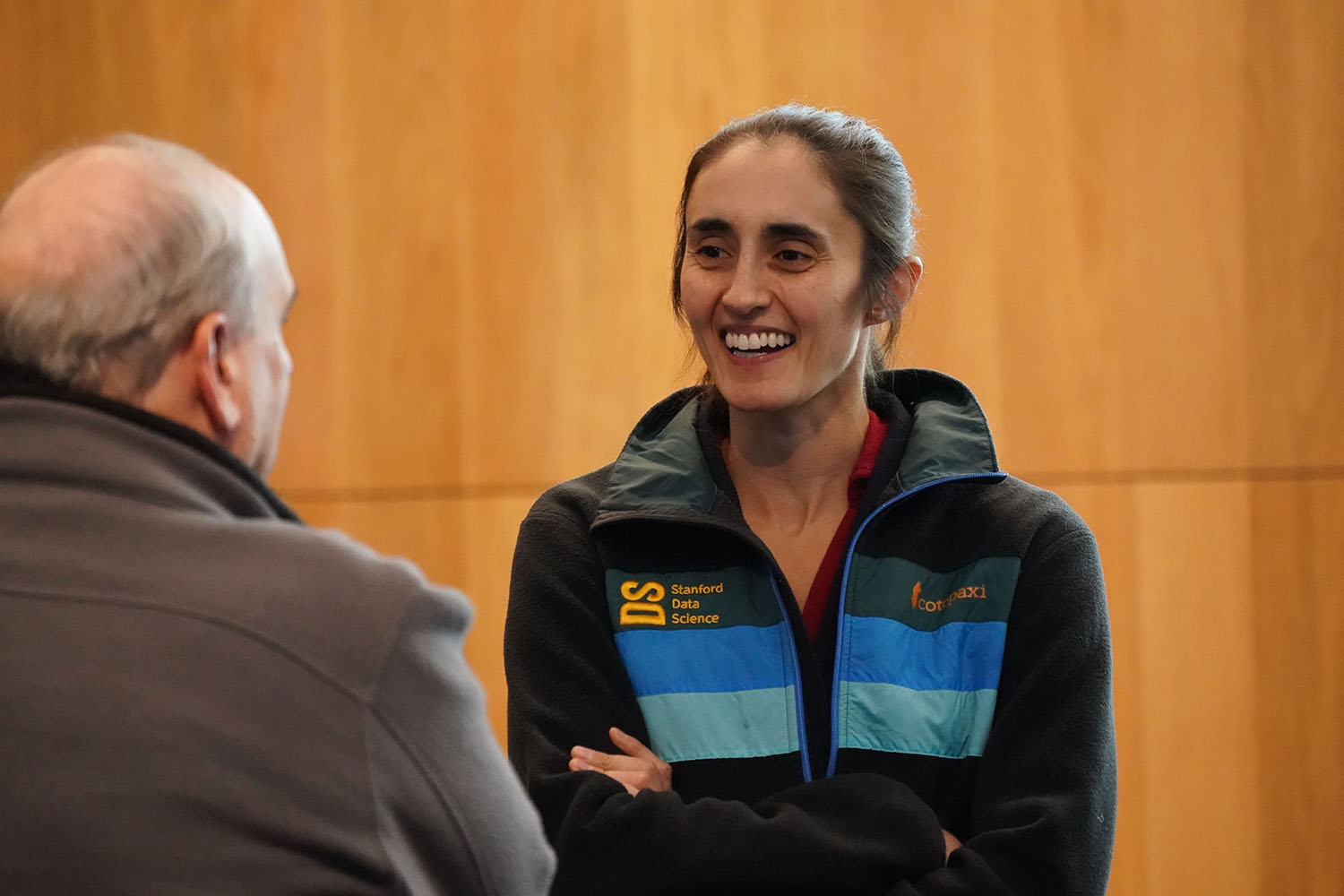 A woman in a Stanford Data Science jacket converses with an older man in front of a wooden backdrop.