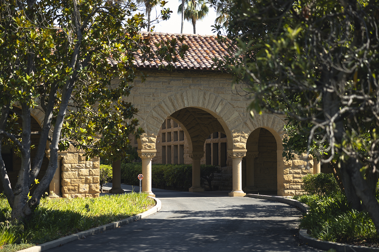 Path through stone archway with red tile roof surrounded by trees on a sunny day.