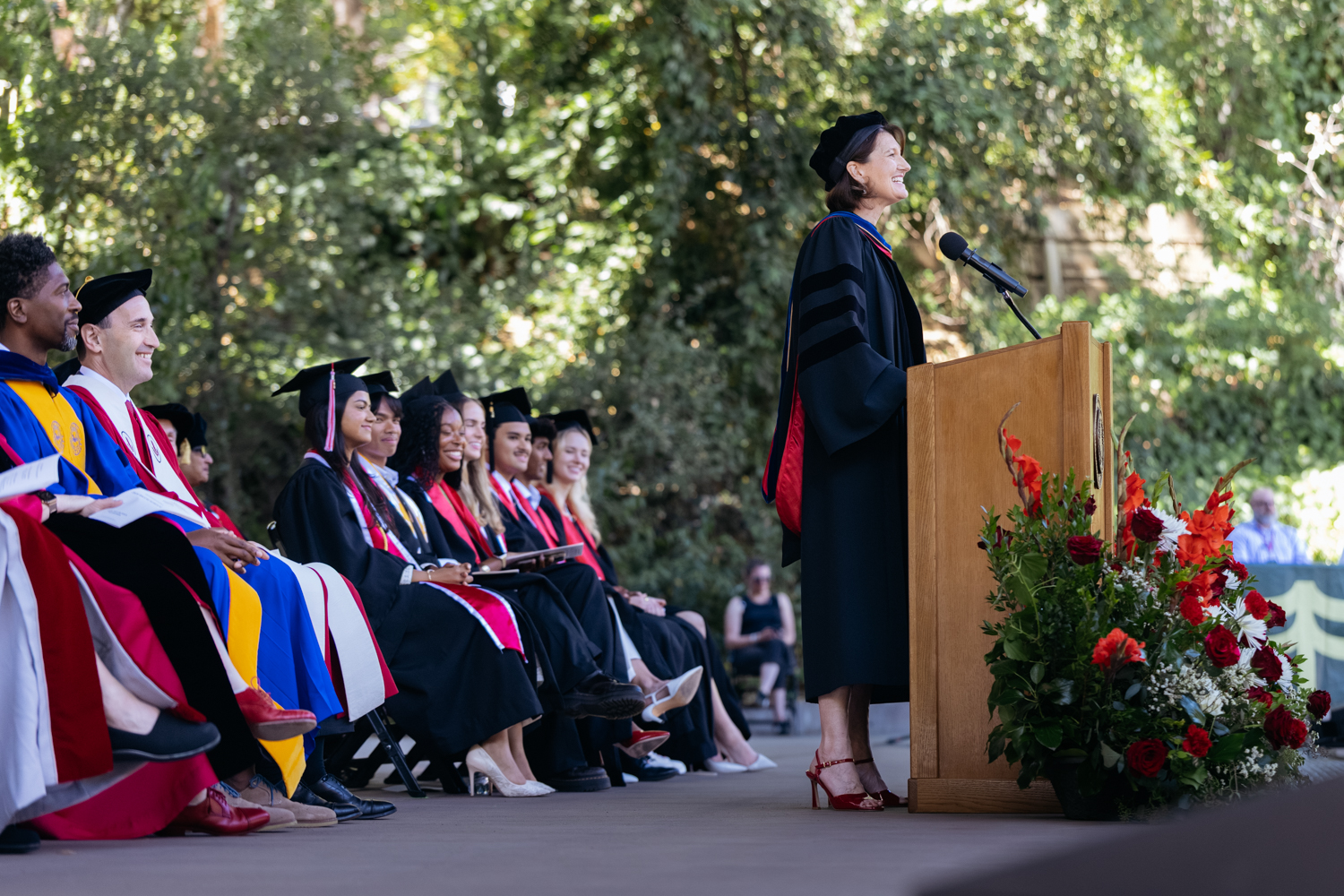 Tiffany Steinwert addresses graduates seated at Baccalaureate, with flowers and green scenery in the background.