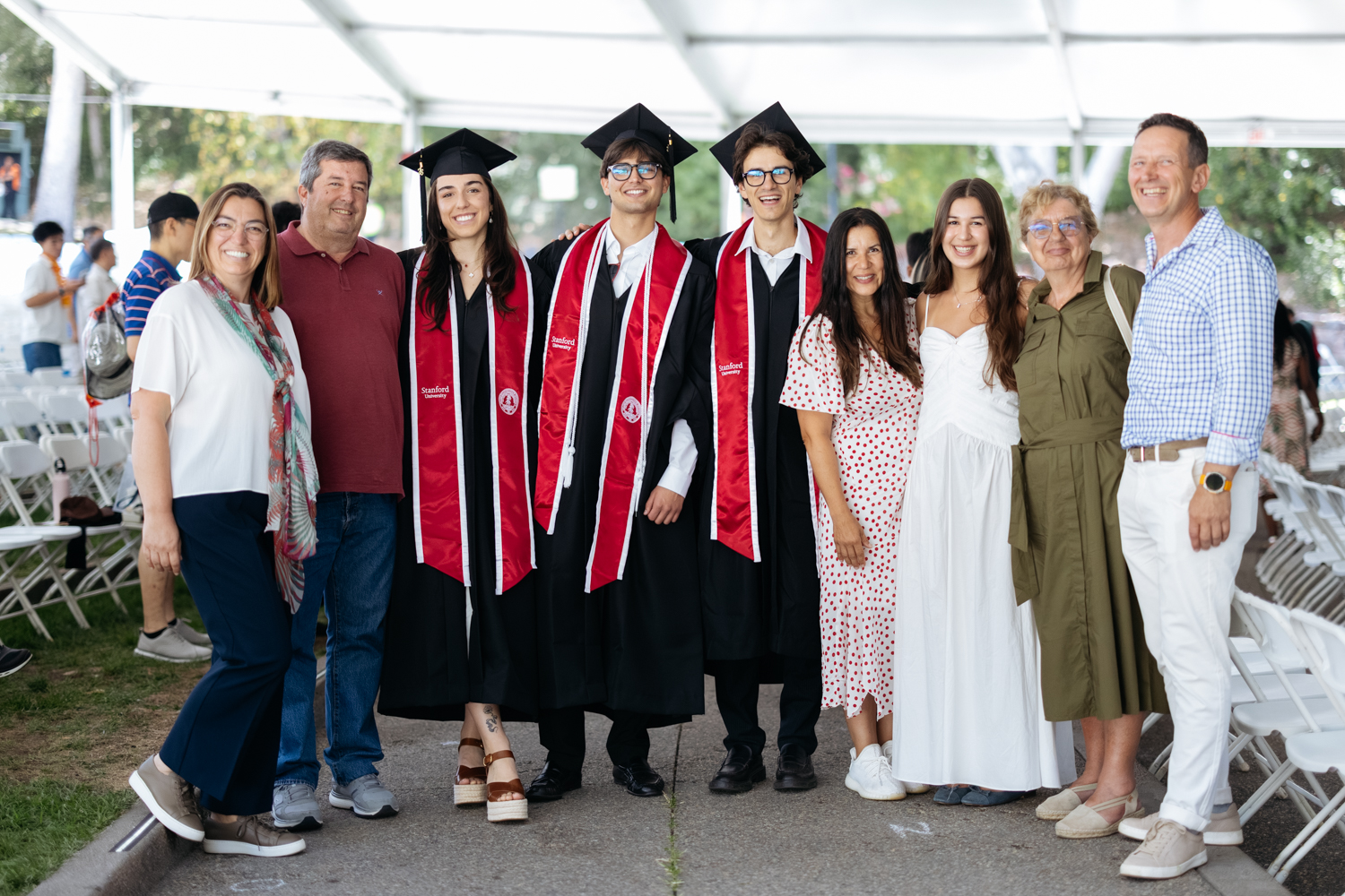 A group of graduates in caps and gowns poses with family members at a graduation ceremony under a tent.