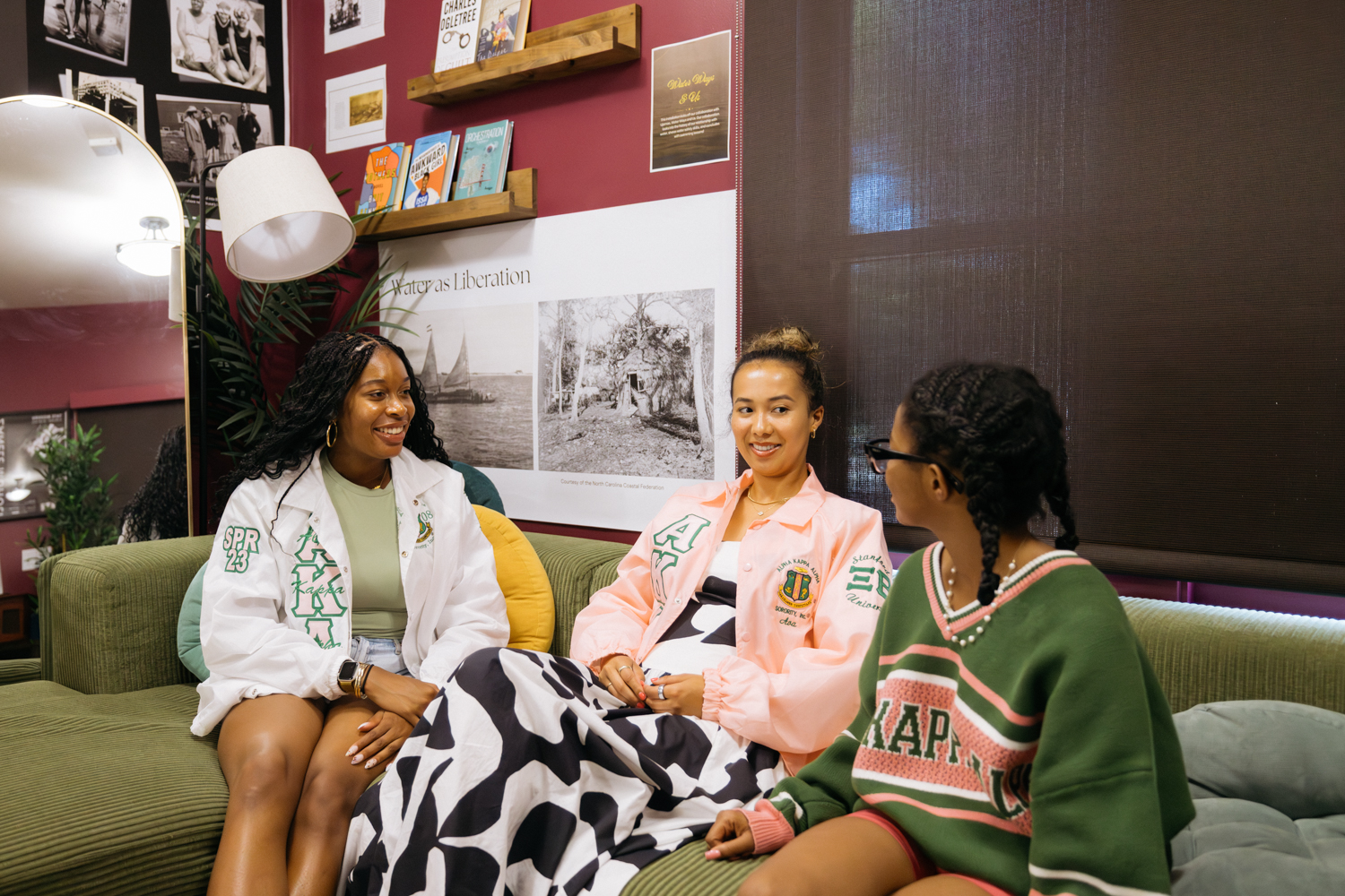 Three people sit together on a couch, engaged in conversation, with artsy decor and historical photos on the walls behind them.