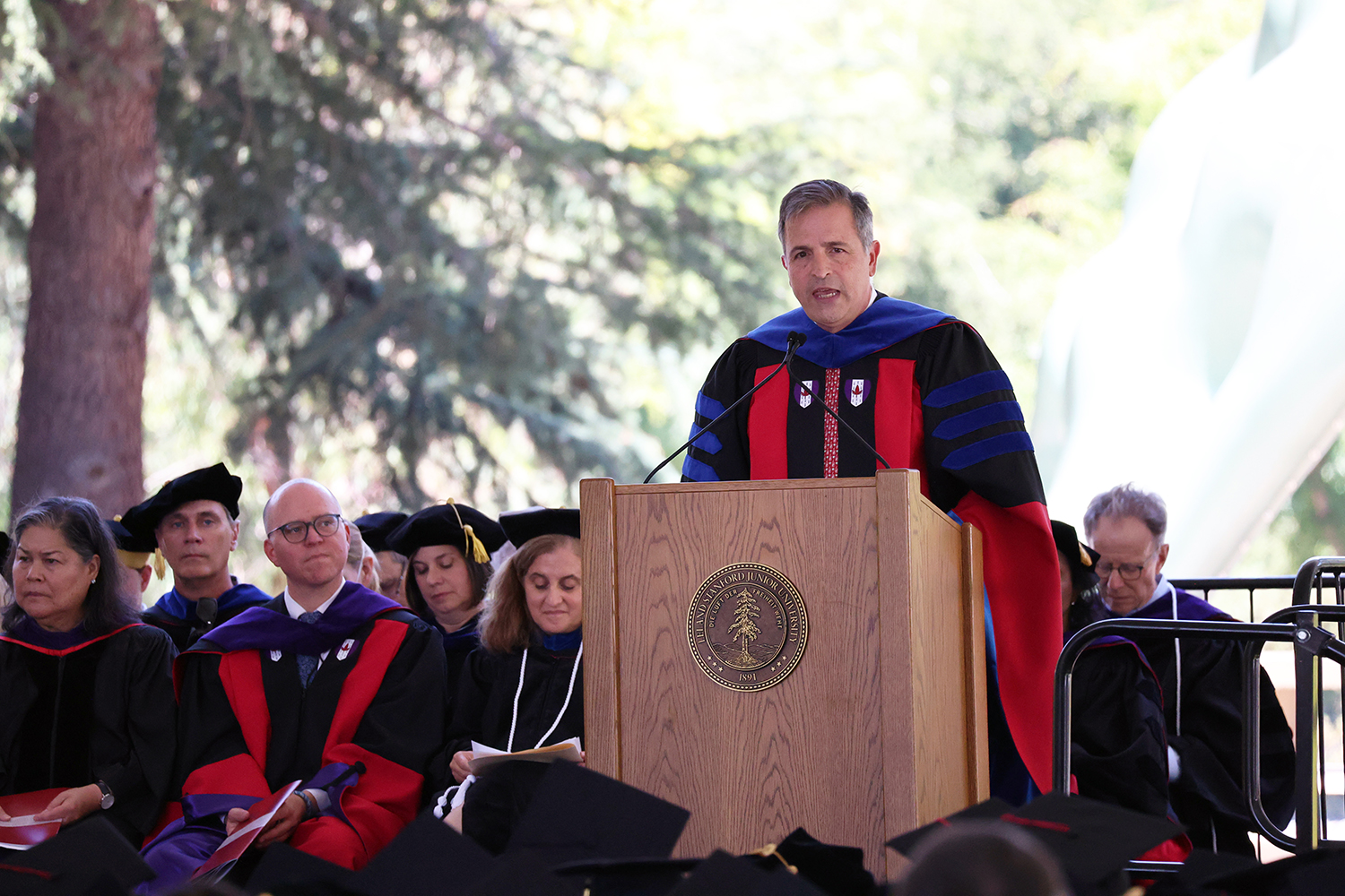 Dean addresses graduates from a podium, with faculty seated attentively under trees at a graduation ceremony.