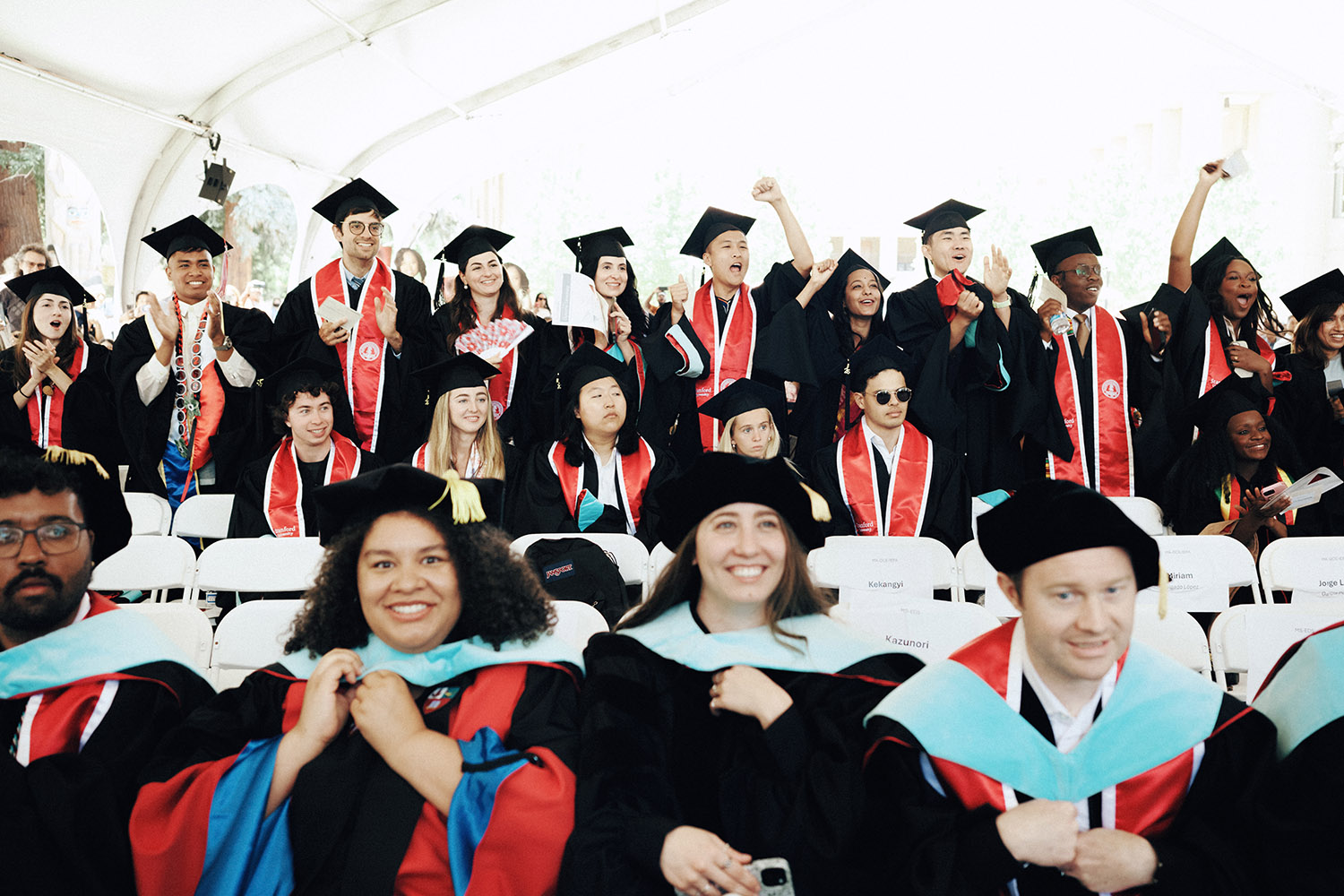 Graduates in caps and gowns celebrate and cheer with raised hands at a graduation ceremony under a tent.