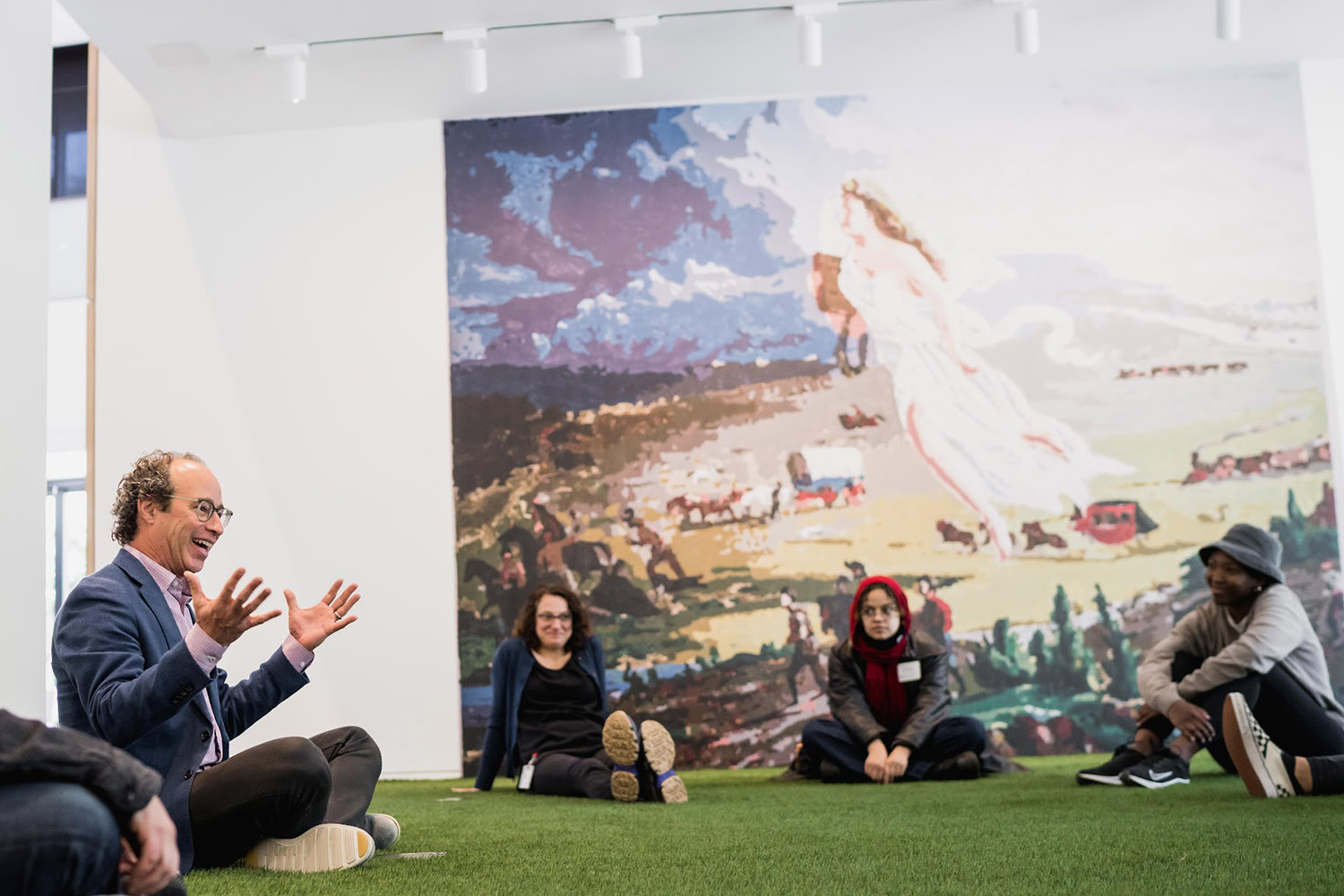 Linetzky gestures while talking to an engaged student audience seated on grass-like flooring in front of a large painting.