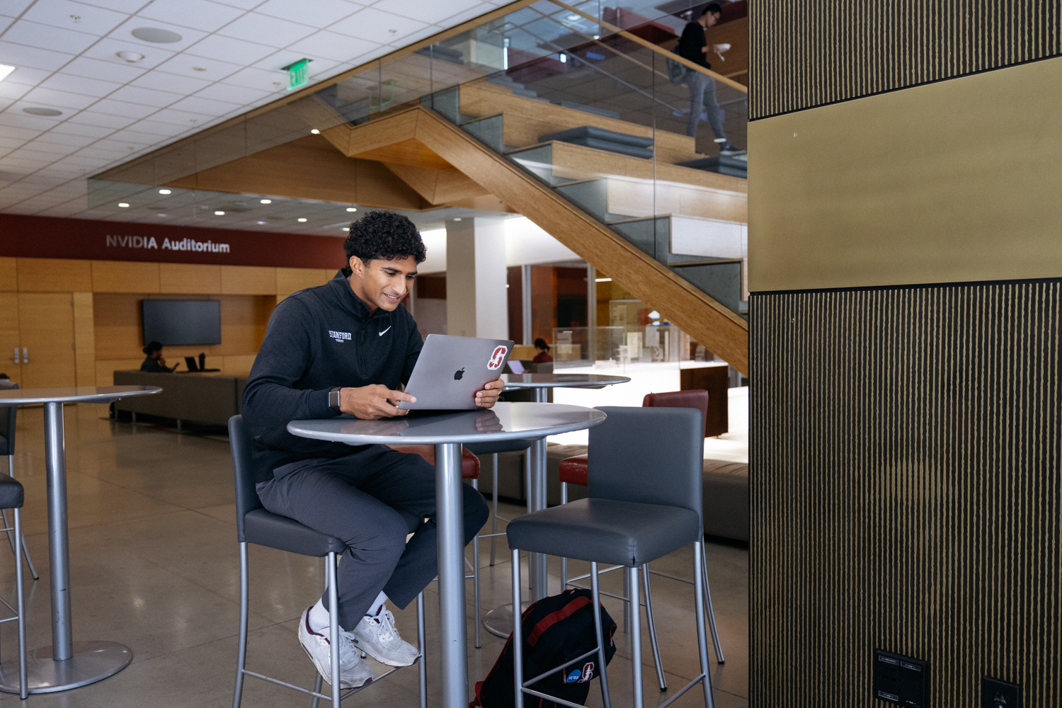 Samir Banerjee studies on a laptop at a high-top table inside the Jen-Hsun Huang Engineering Center, with the NVIDIA Auditorium entrance in the background.