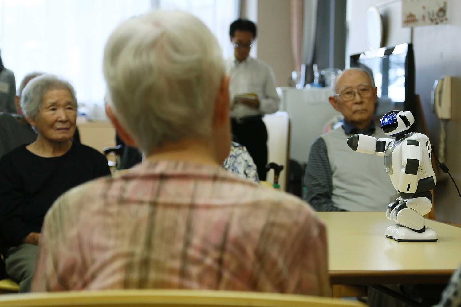 A small robot interacts with elderly people in a care setting, while staff assist in the background.