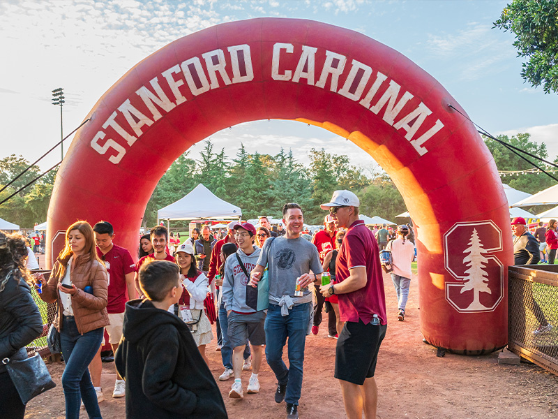 Fans walk through an inflatable arch reading Stanford Cardinal at a tailgate before a game.