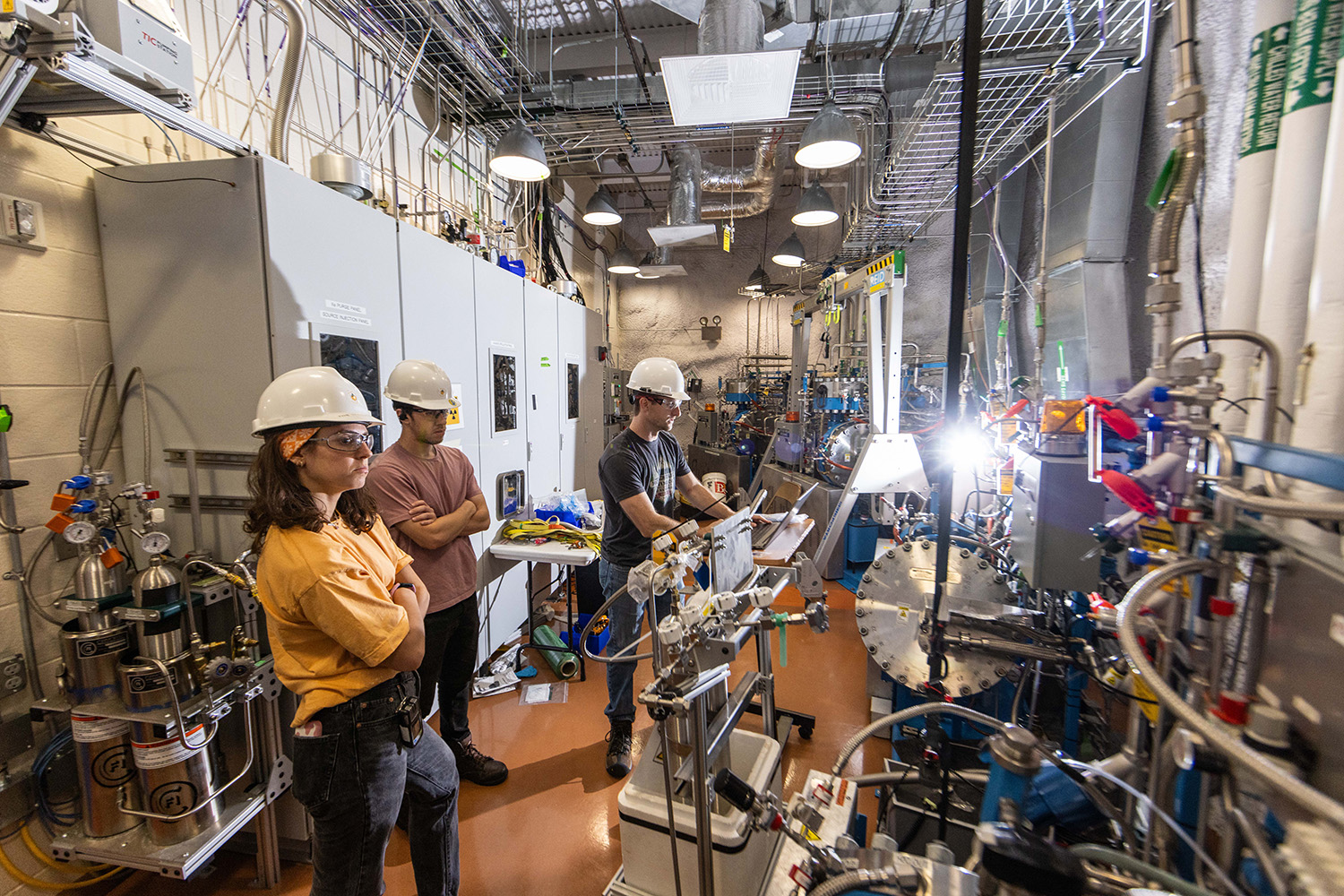 Three researchers in hard hats monitor equipment in a crowded lab filled with pipes, valves, and control panels.