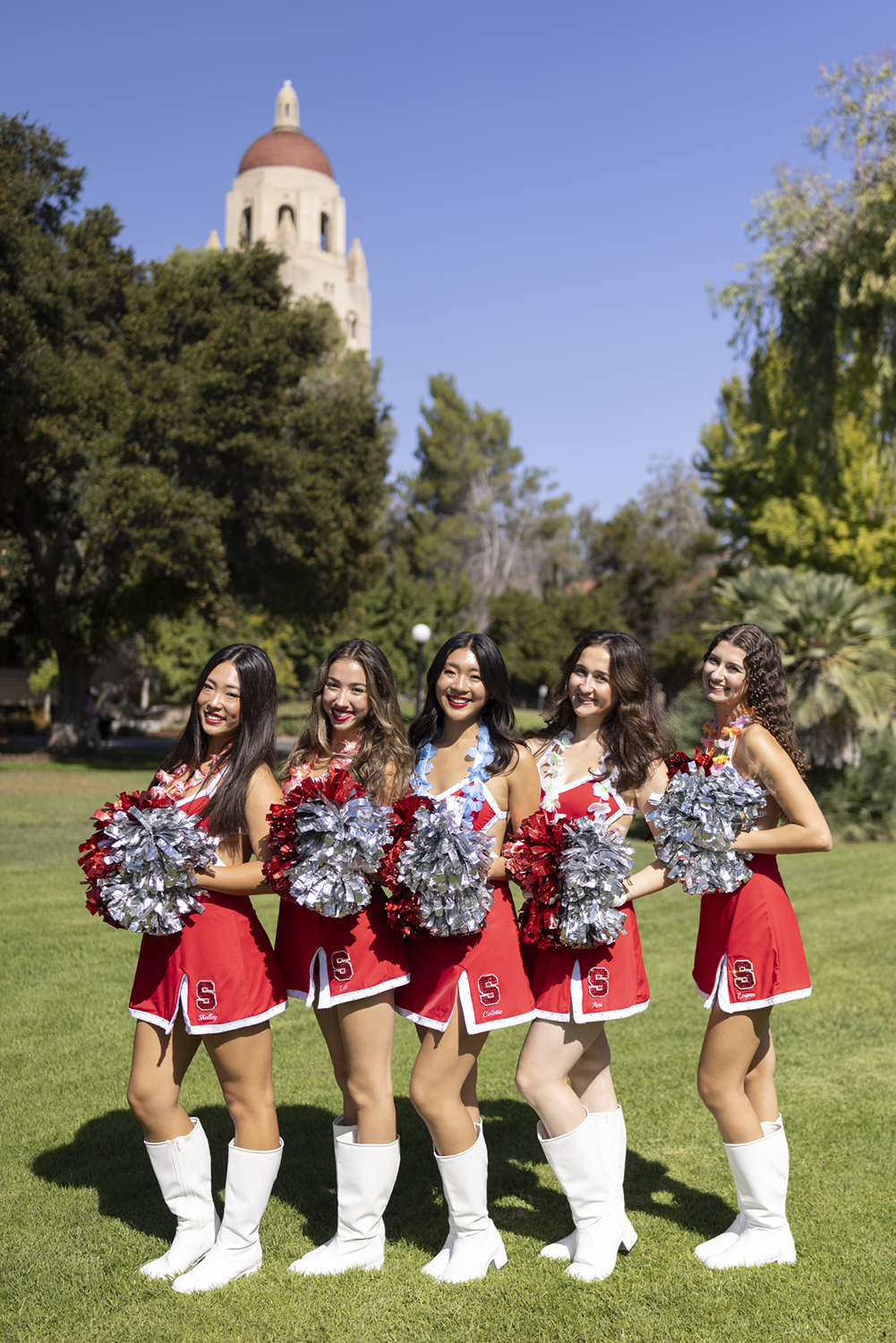 Five cheerleaders in red uniforms and pom-poms pose on the lawn with Hoover Tower in the background.