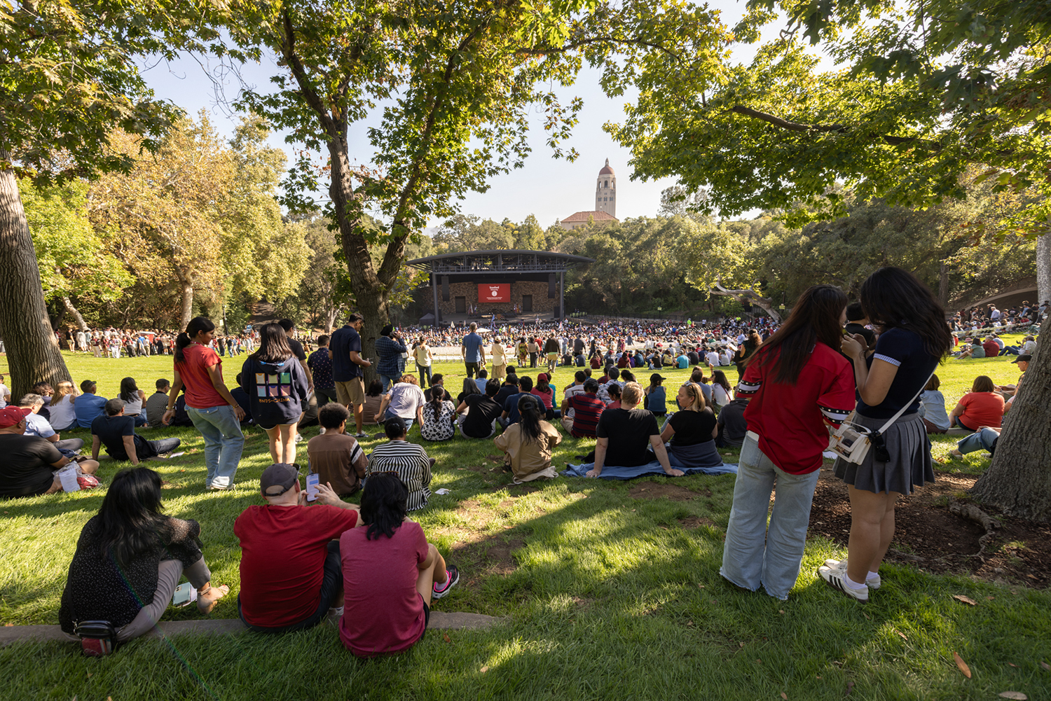 Families watch the Opening Convocation Ceremony from a shaded lawn as a large screen shows the stage in the distance.
