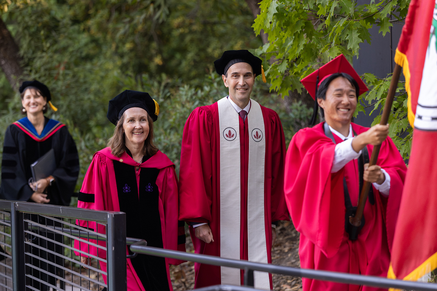 University leaders in academic robes walk together outdoors, with one carrying a ceremonial flag.