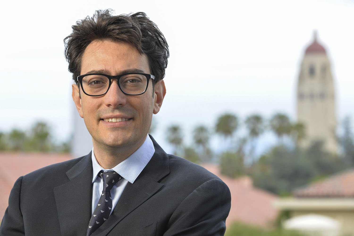 Marco Pavone in dark suit and glasses smiling outdoors, with Hoover Tower blurred in the background.