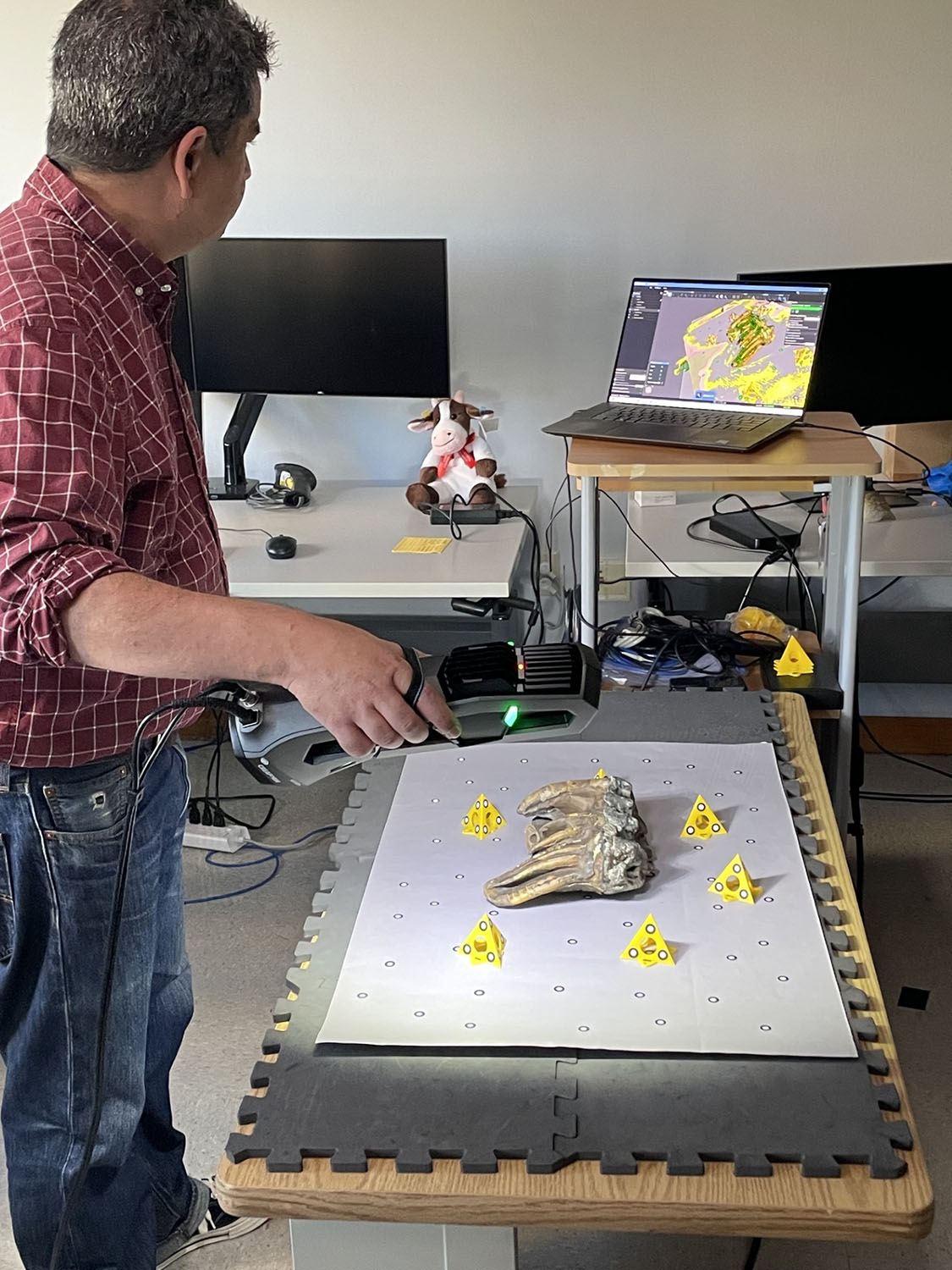 Tony Calavano holds handheld 3D scanner over fossil tooth on a scanning table; laptop shows colored scan model.
