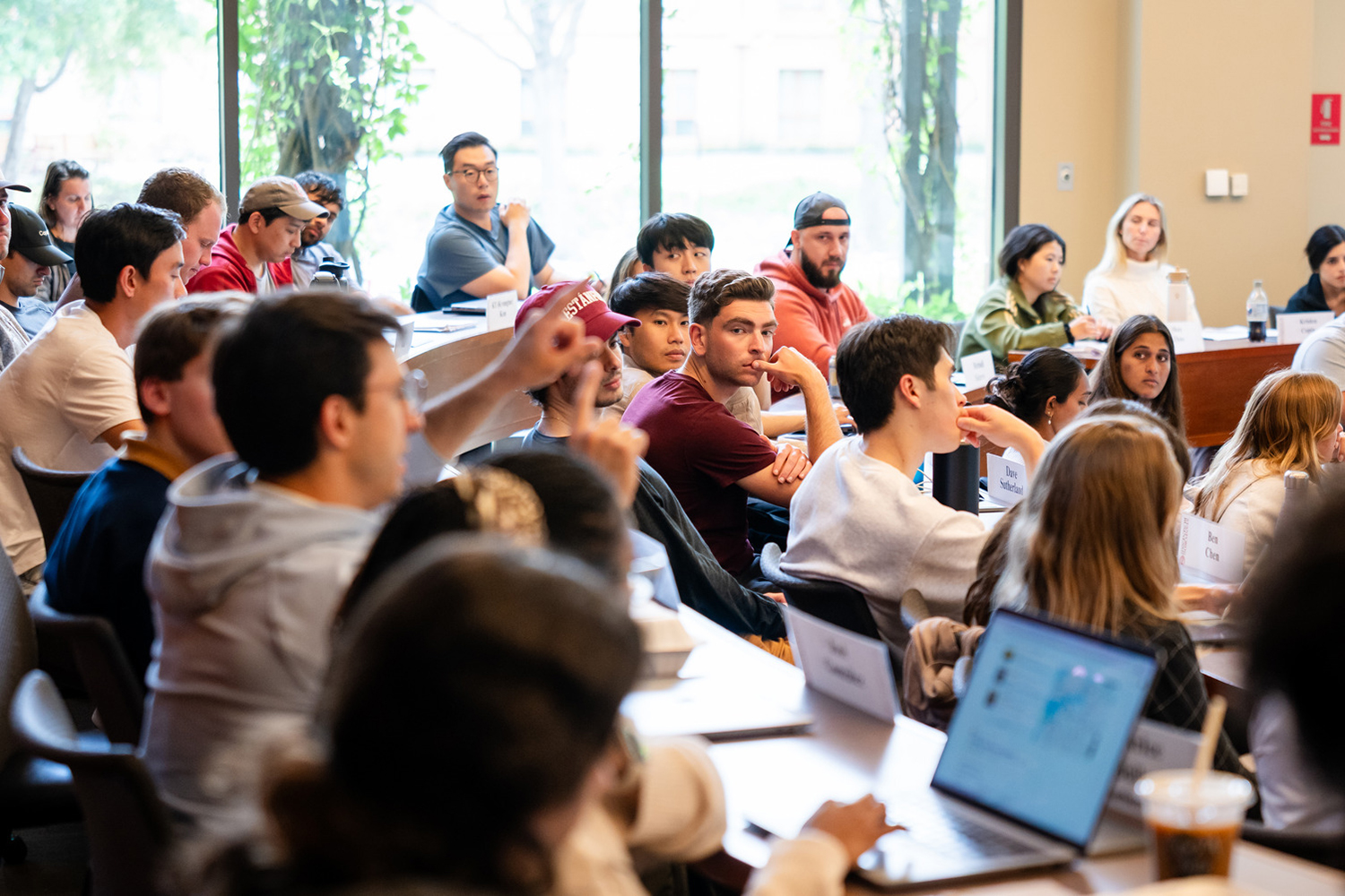 Business school students sit at name-labeled desks in a discussion-style classroom as a peer raises his hand.
