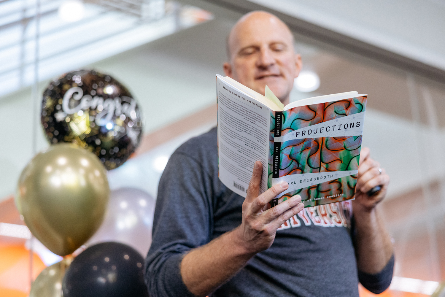 Markus Covert reads aloud from an open copy of the book Projections beside gold, black, and silver balloons.