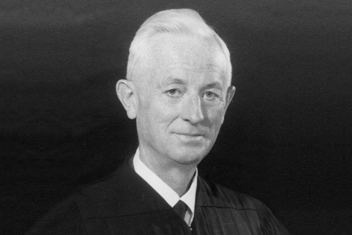 Judge Duniway, a smiling, white-haired man in a judicial robe and tie against a dark background.