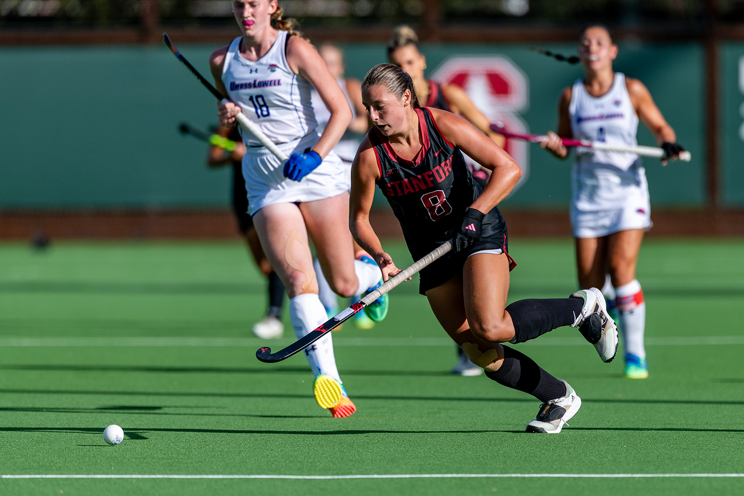 A Cardinal field hockey player dribbles the ball with her stick as UMass Lowell defenders chase behind her.
