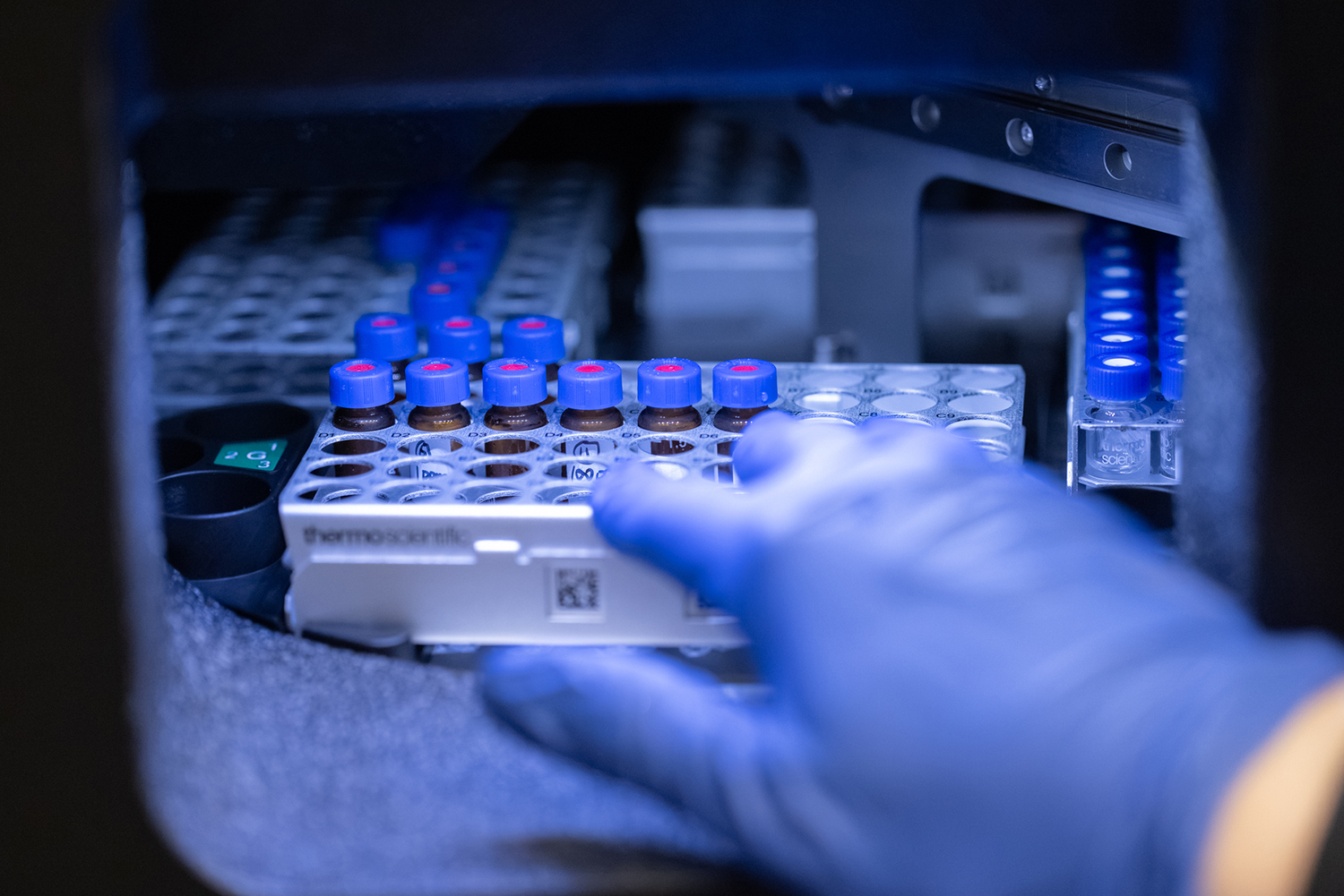 A gloved hand rests on a rack of capped sample vials inside a laboratory instrument.