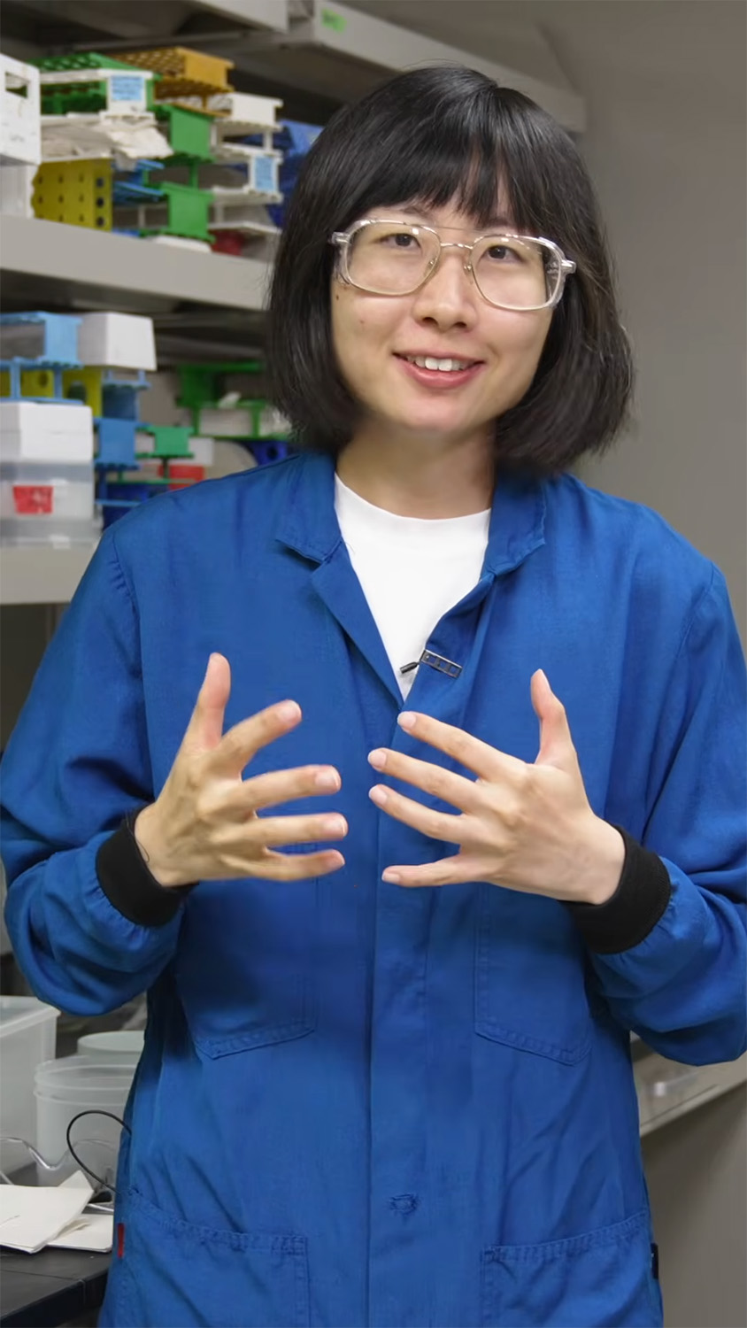 Nina Zhao speaking and gesturing with both hands during an interview in front of lab shelves stocked with sample racks.