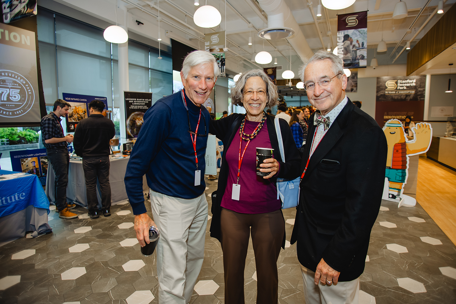 Three smiling attendees wearing lanyards pose together in a busy hall lined with exhibitor tables and anniversary banners.