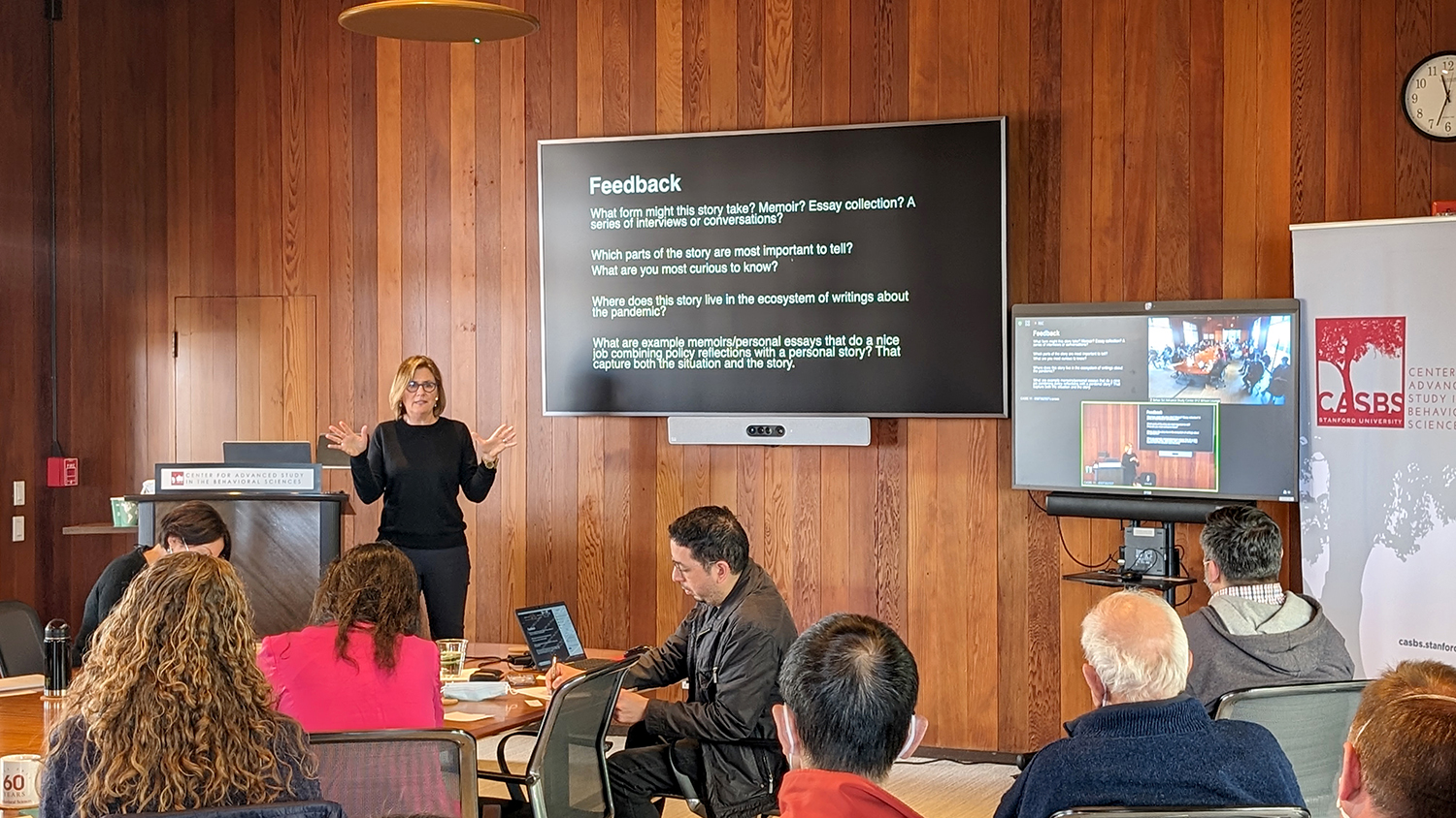 Dr. Sara Cody standing at the front of a room next to a podium and powerpoint presentation and is speaking to a small audience seated around a variety of tables.