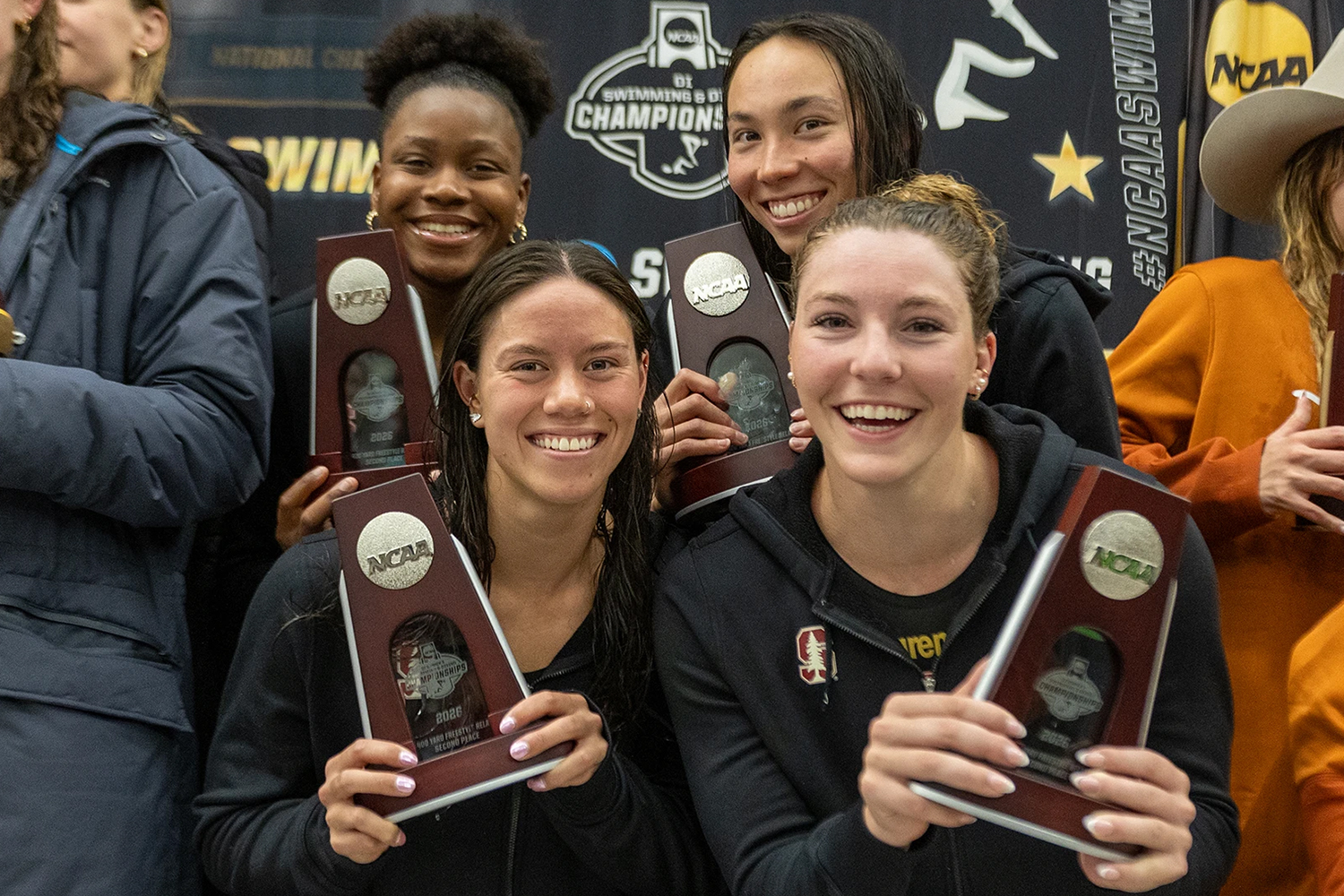 Four smiling swimmers pose together holding NCAA championship trophies in front of a branded backdrop.