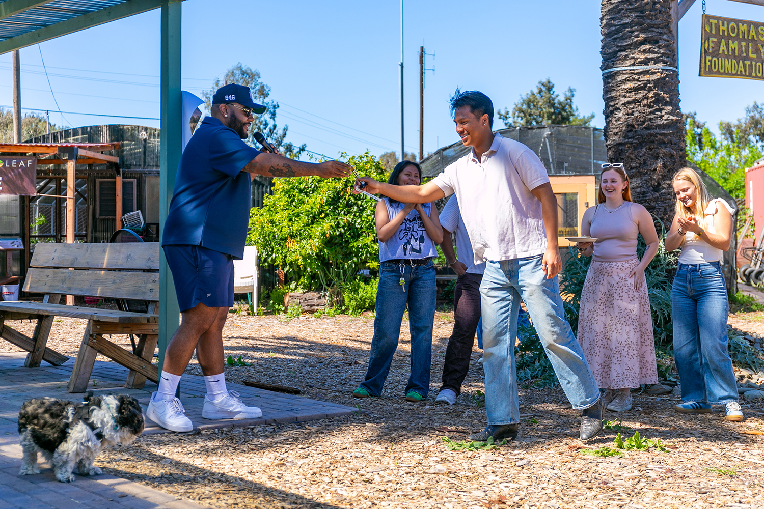 Ejaaz hands out an end-of-course award to a student while a small dog and several people observe in the background.