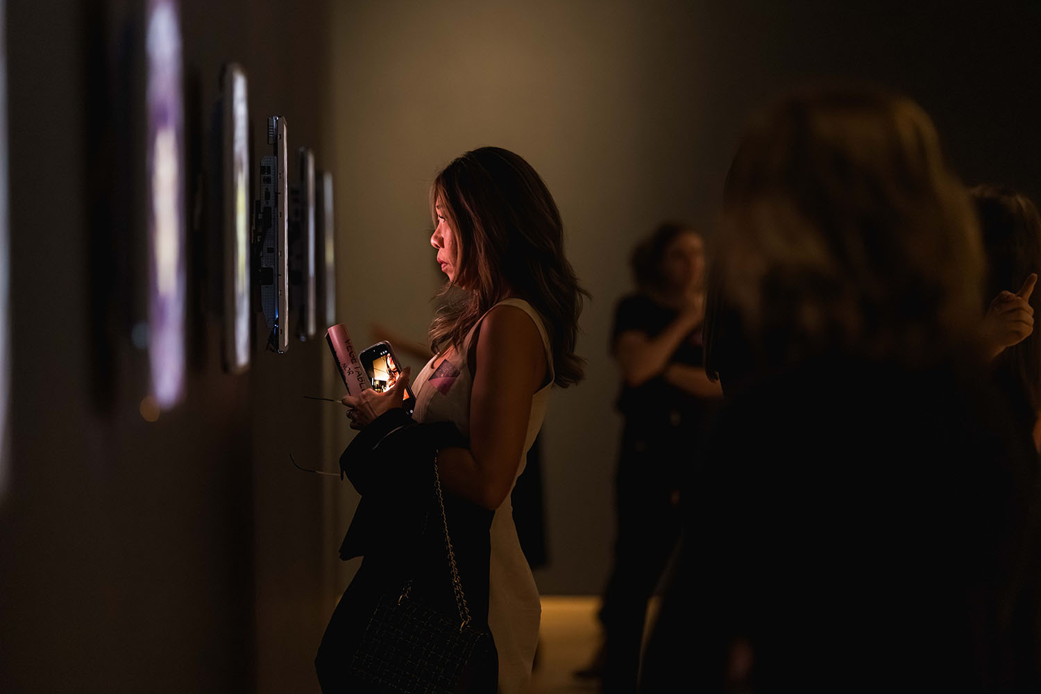 Visitor holding phone views artwork in a dimly lit gallery.
