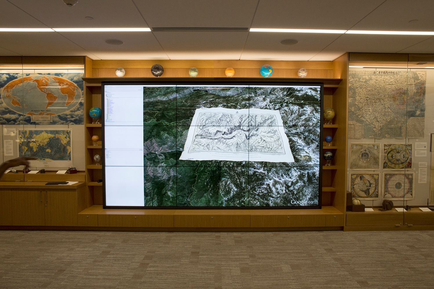 A large display shows a 3D map overlay of mountainous terrain, surrounded by educational maps and globes in a learning environment.