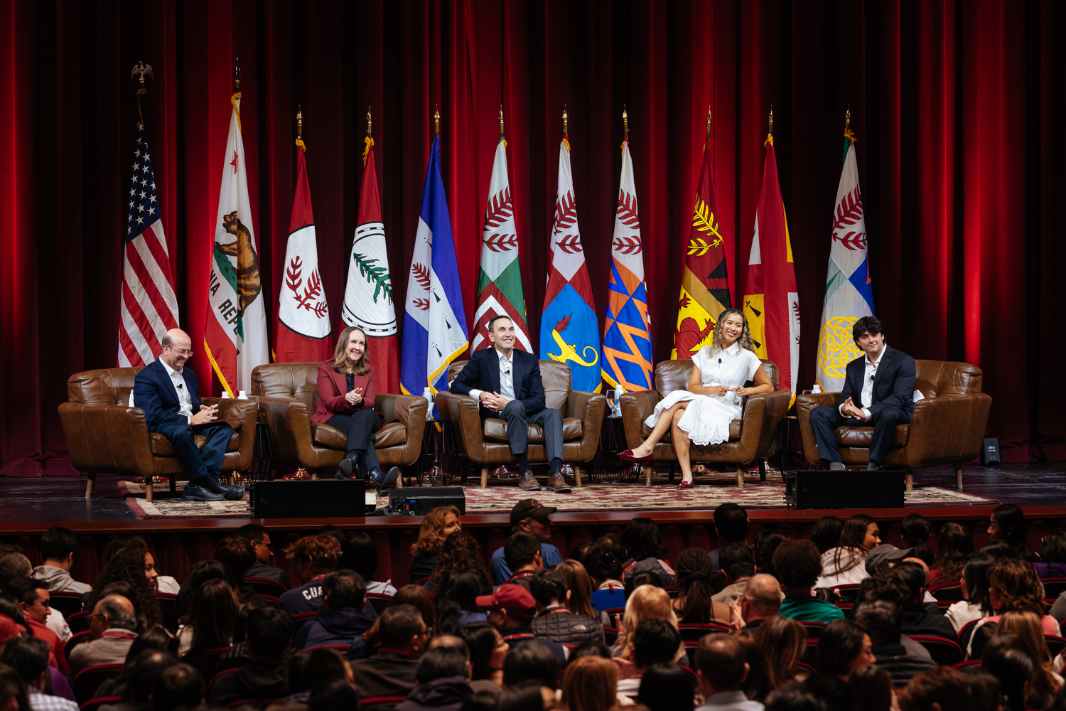 A panel discussion with four speakers seated on couches, facing an audience, with multiple flags displayed in the background.