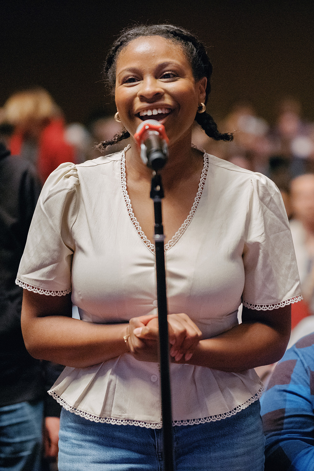 A person in a light blouse stands at a microphone, with hands clasped, in front of an audience at an event.