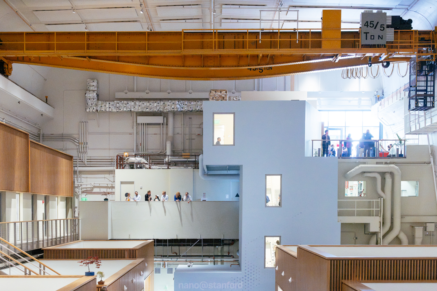 A spacious industrial setting features a yellow crane overhead, with people observing from a balcony and a modern architectural design below.