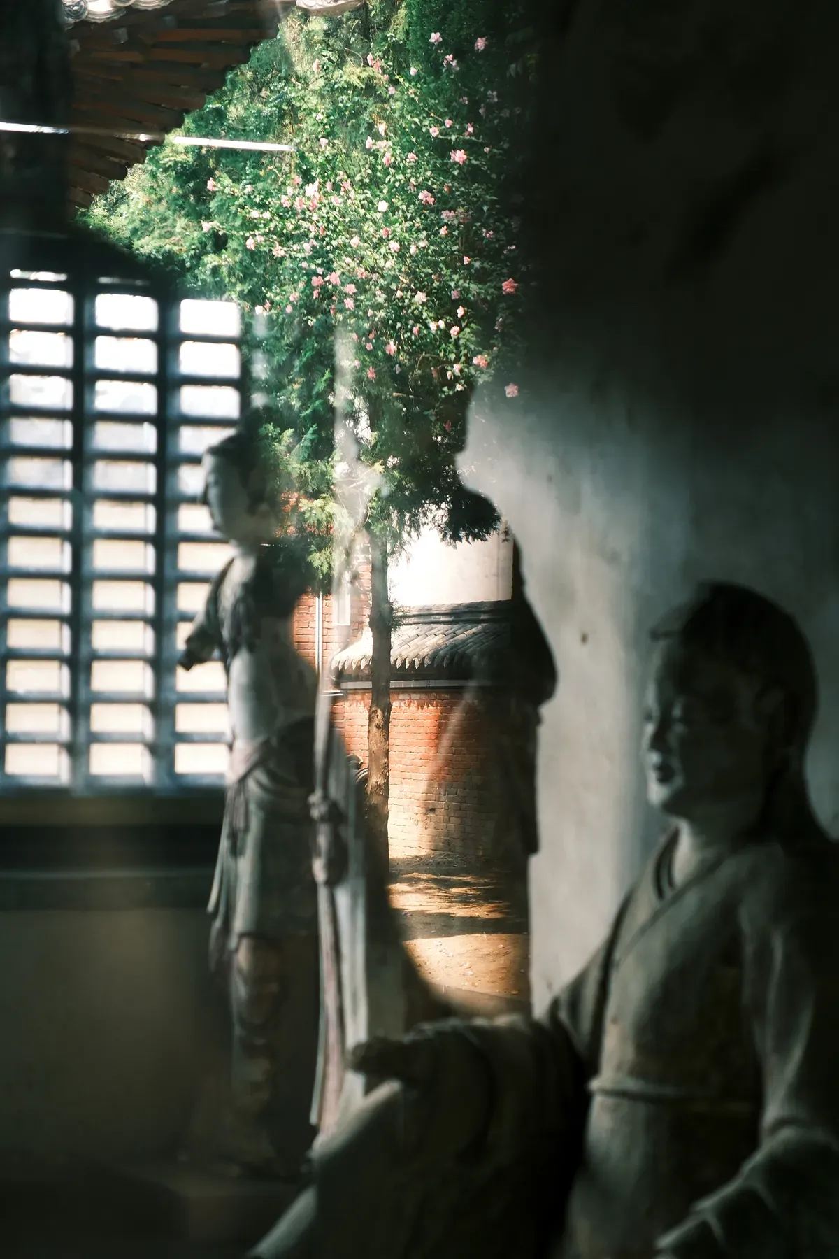 Statues are reflected in a window showing a garden with blooming flowers and a glimpse of a traditional building outside.