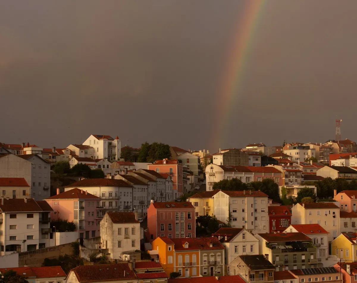 A view of colorful buildings in a cityscape under a cloudy sky, featuring a faint rainbow arching over the rooftops.