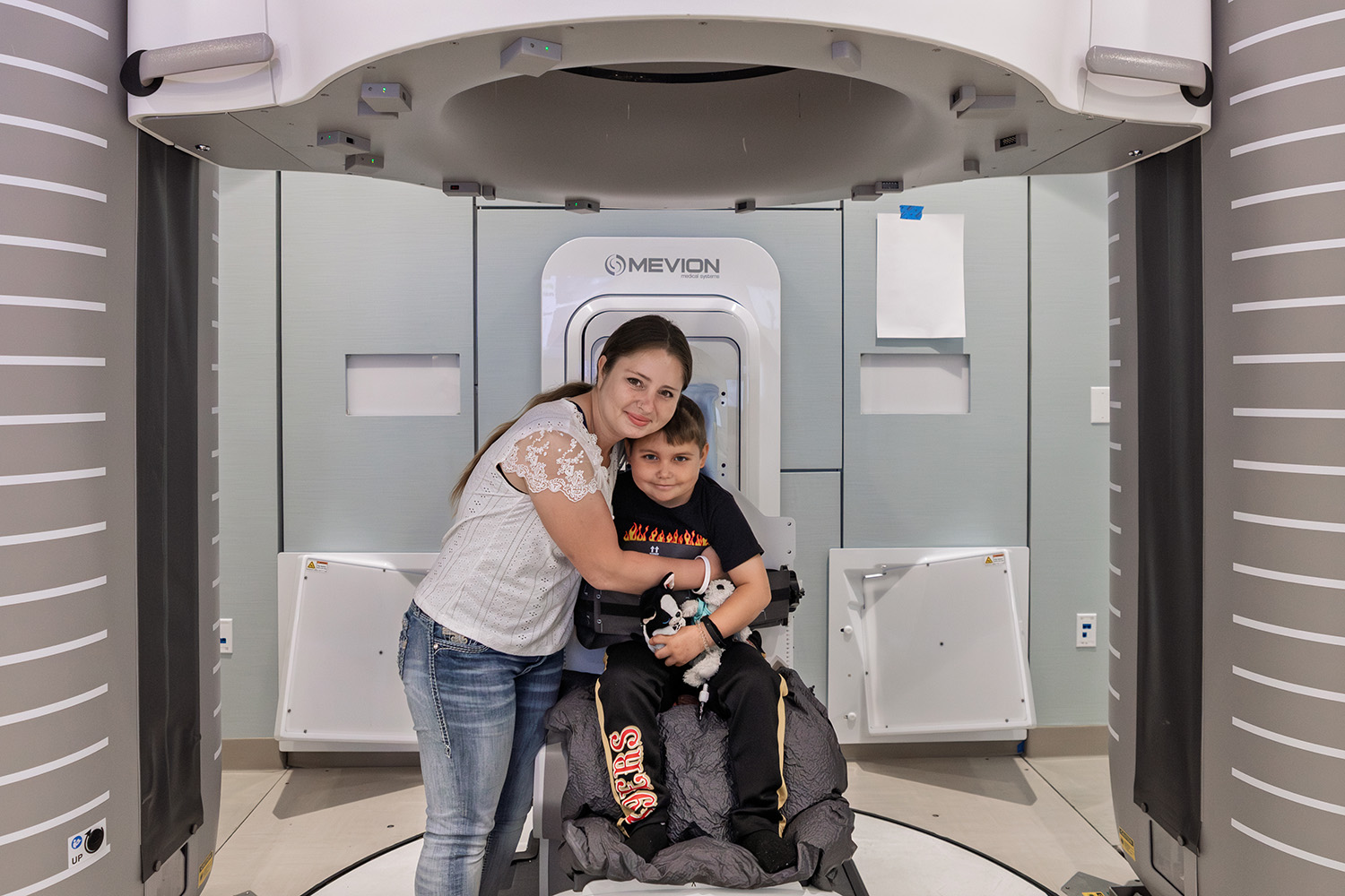A child sits in a medical chair inside a machine while his mother hugs him, preparing for a treatment procedure.