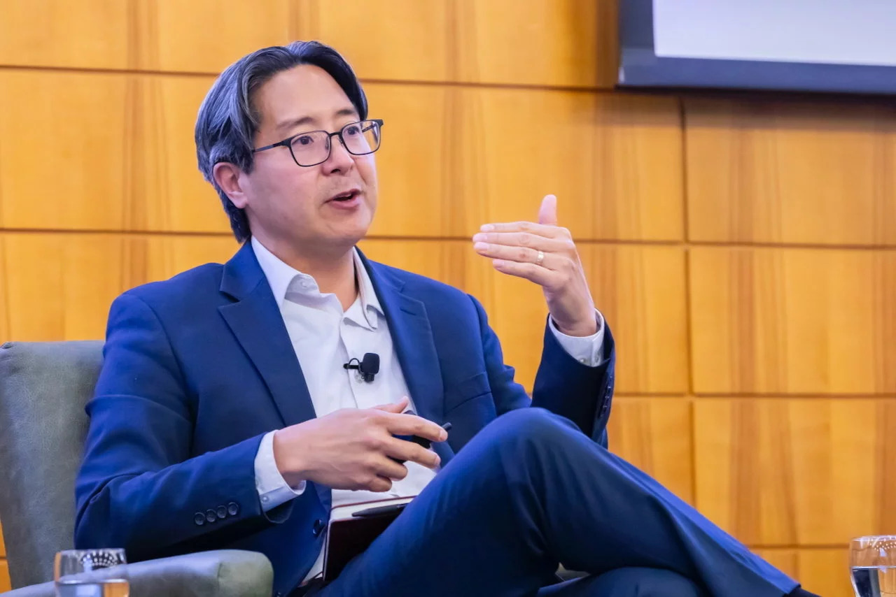 Open video Michael Hsu in a suit speaks while seated in a chair, gesturing with one hand during a panel discussion. in a modal