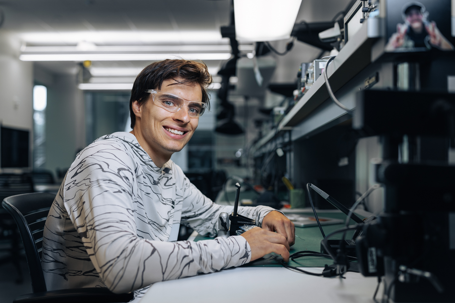 Connor Herson in a patterned hoodie works intently at a cluttered desk with electronic equipment in a tech lab setting.