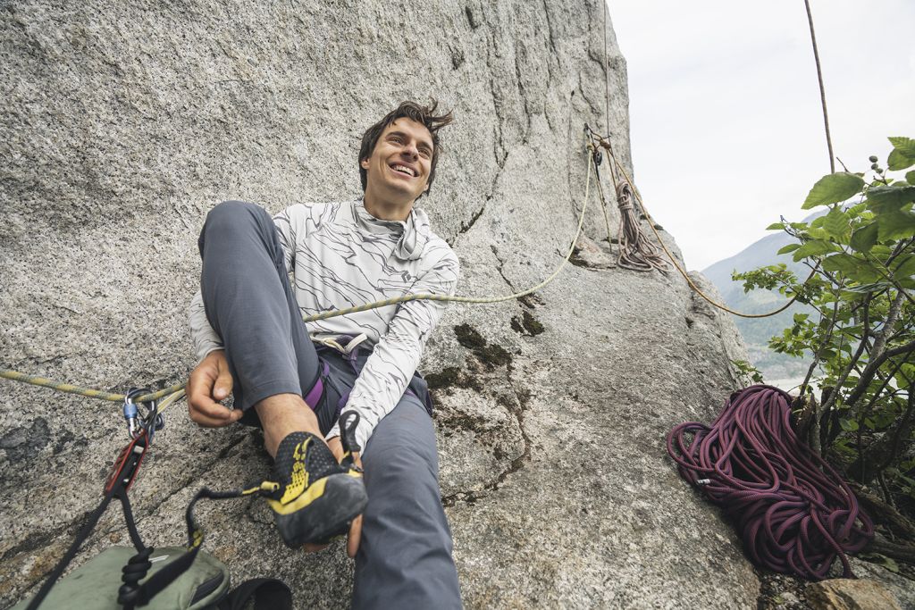 Connor Herson sits on a rocky surface, adjusting their climbing shoe and harness, with climbing ropes visible nearby.
