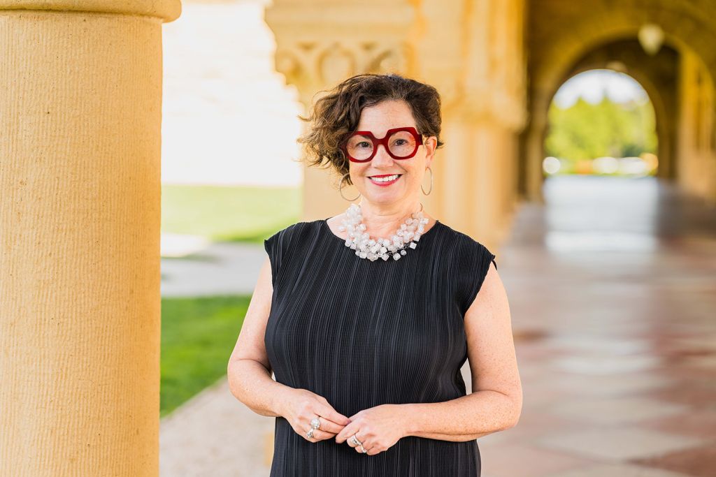 Jennifer Devere Brody stands near a column, smiling for a profile photo in a sunny outdoor setting with arches in the background.